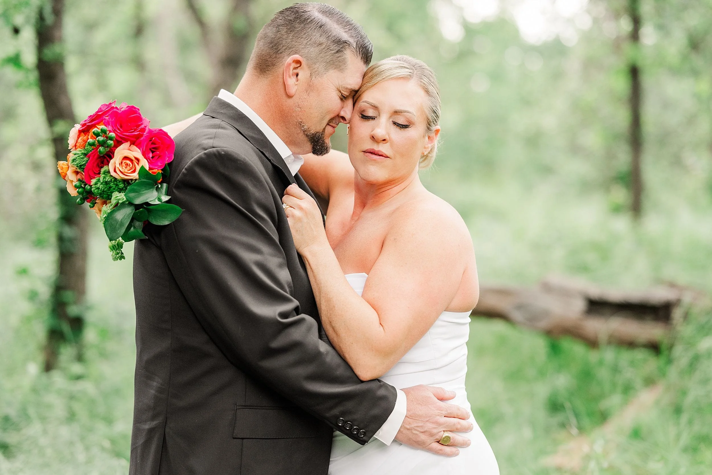A couple embraces tenderly in a lush, green forest. The woman in a white dress holds a vibrant bouquet of pink and orange flowers. The scene is serene and romantic.