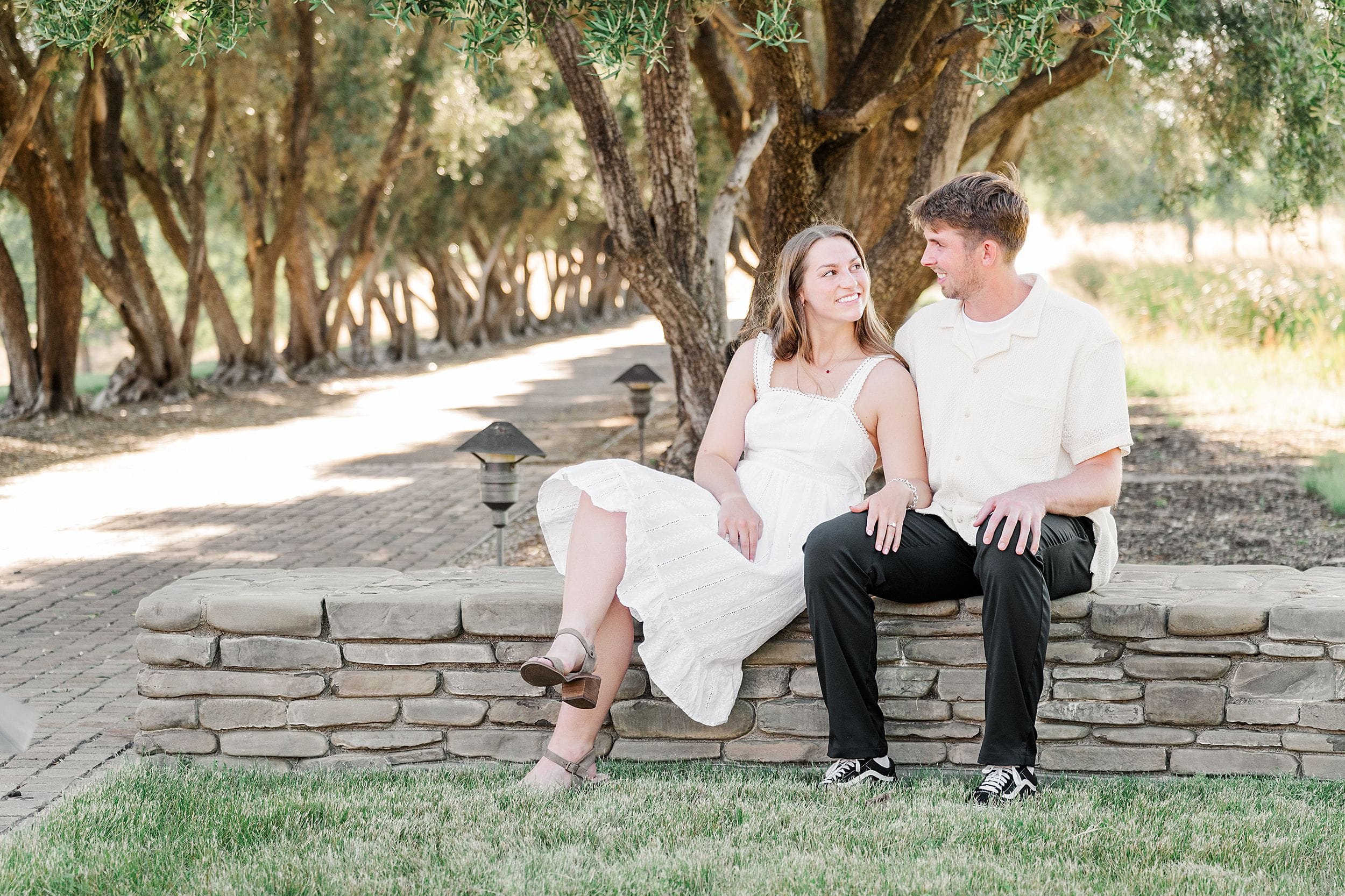 A woman in a white dress and a man in a light shirt and black pants sit on a stone wall under trees, looking at each other and smiling.