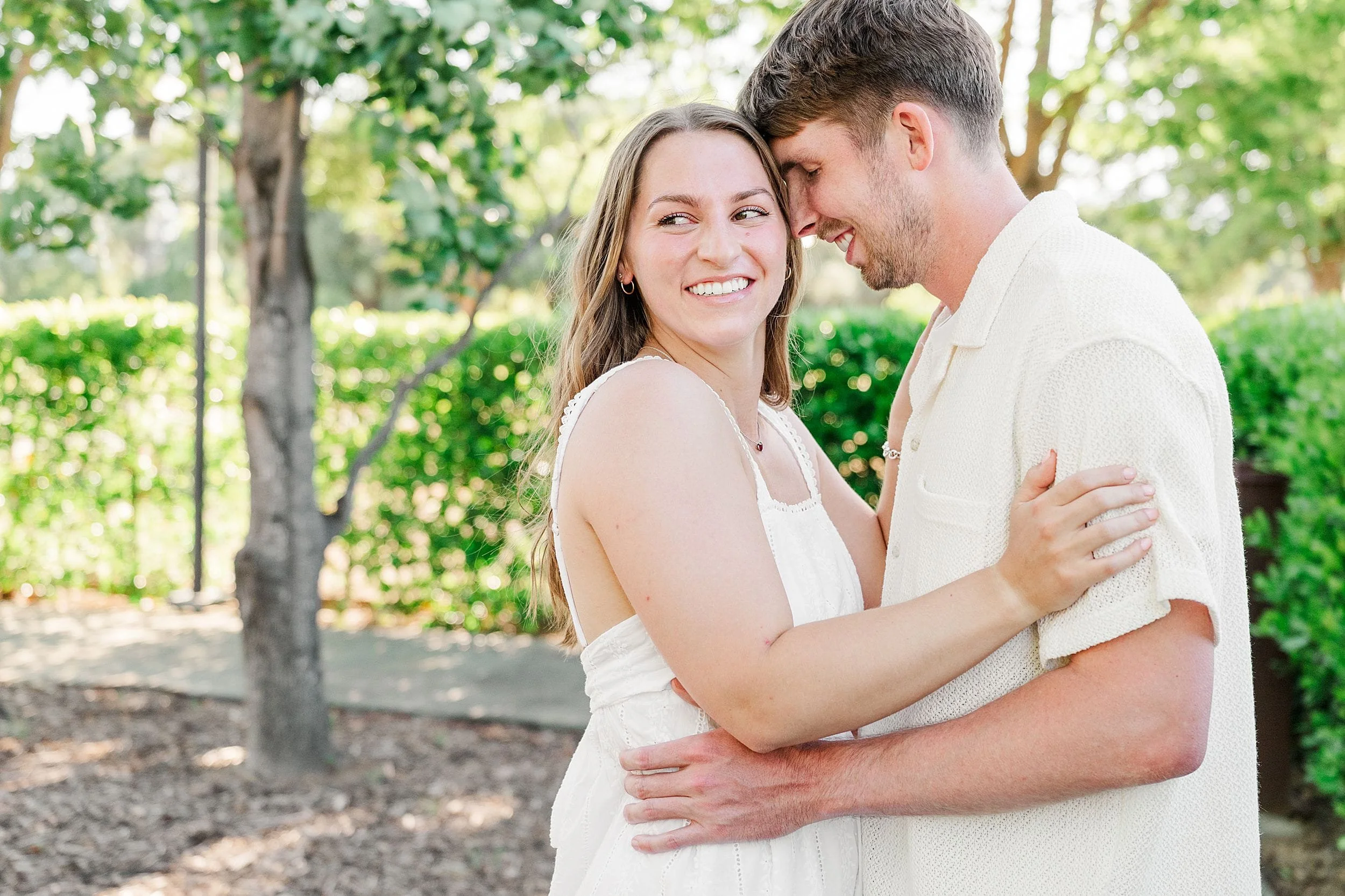 A smiling couple embraces in a sunlit garden. Both wear white outfits, conveying joy and affection. Lush greenery surrounds them, enhancing the warm atmosphere.