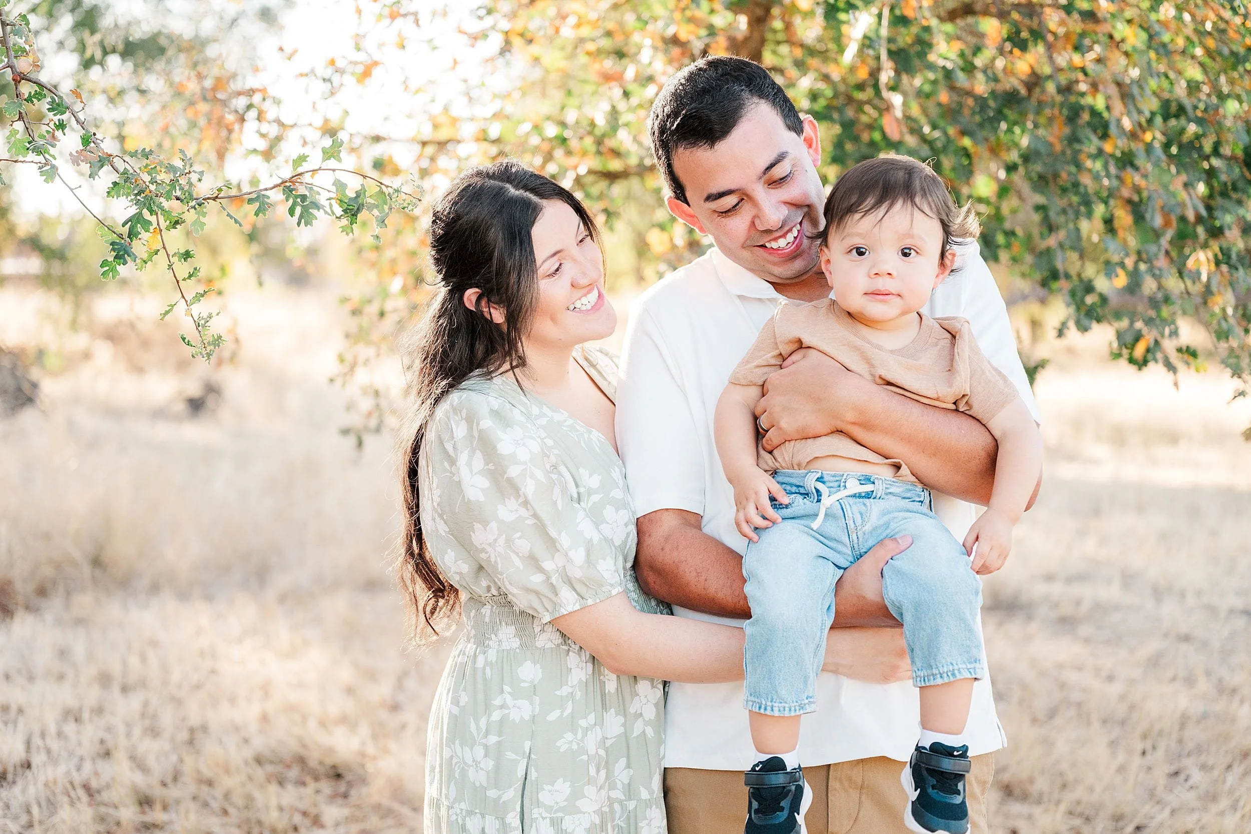 A smiling couple holds their toddler outdoors beneath golden autumn leaves. The warm, sunlit family portrait conveys affection, joy, and togetherness.