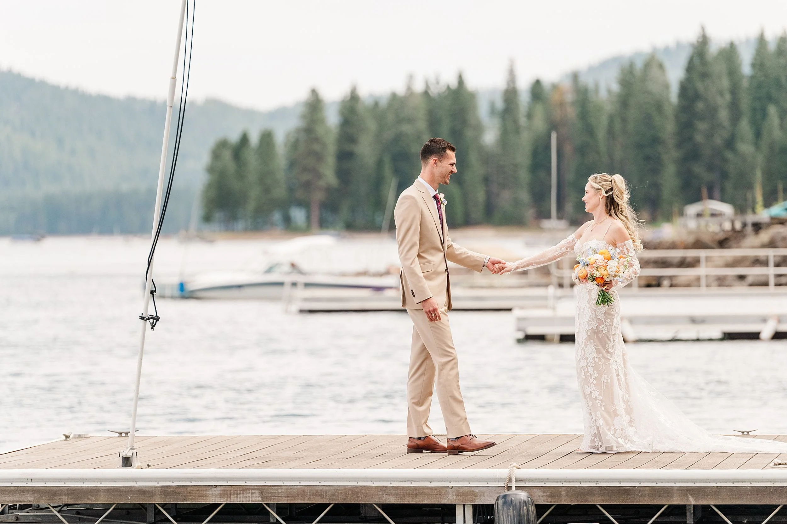 Bride and groom hold hands on a lakeside dock, smiling in wedding attire, with sailboats, pine forest, and mountains creating a romantic scene.
