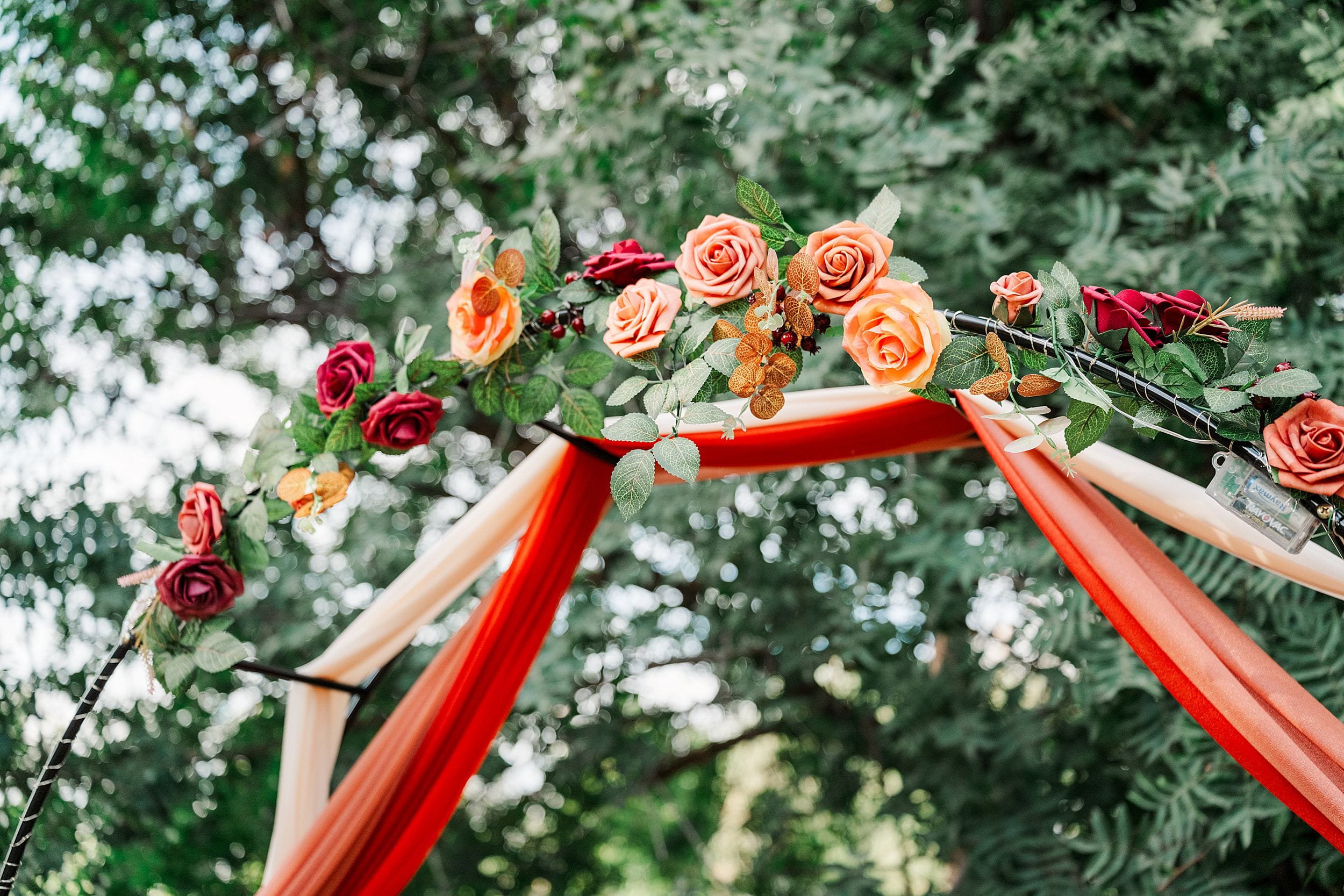 Outdoor wedding arch draped in red and peach fabric, adorned with roses and greenery, creating a romantic, festive setting beneath trees.