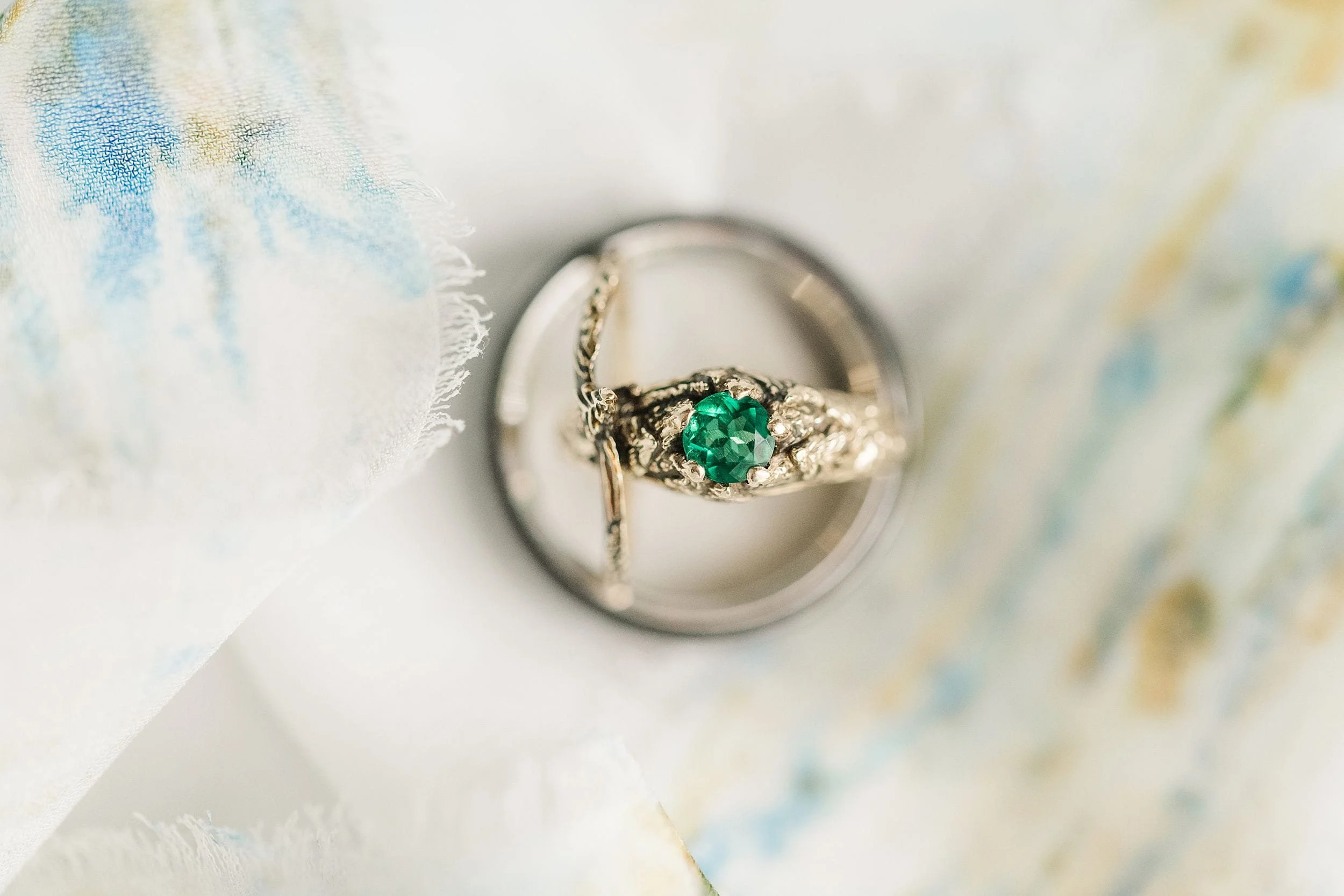 An ornate gold ring with a vibrant green gemstone rests atop a silver band on a soft, pastel-colored fabric, suggesting elegance and romance.
