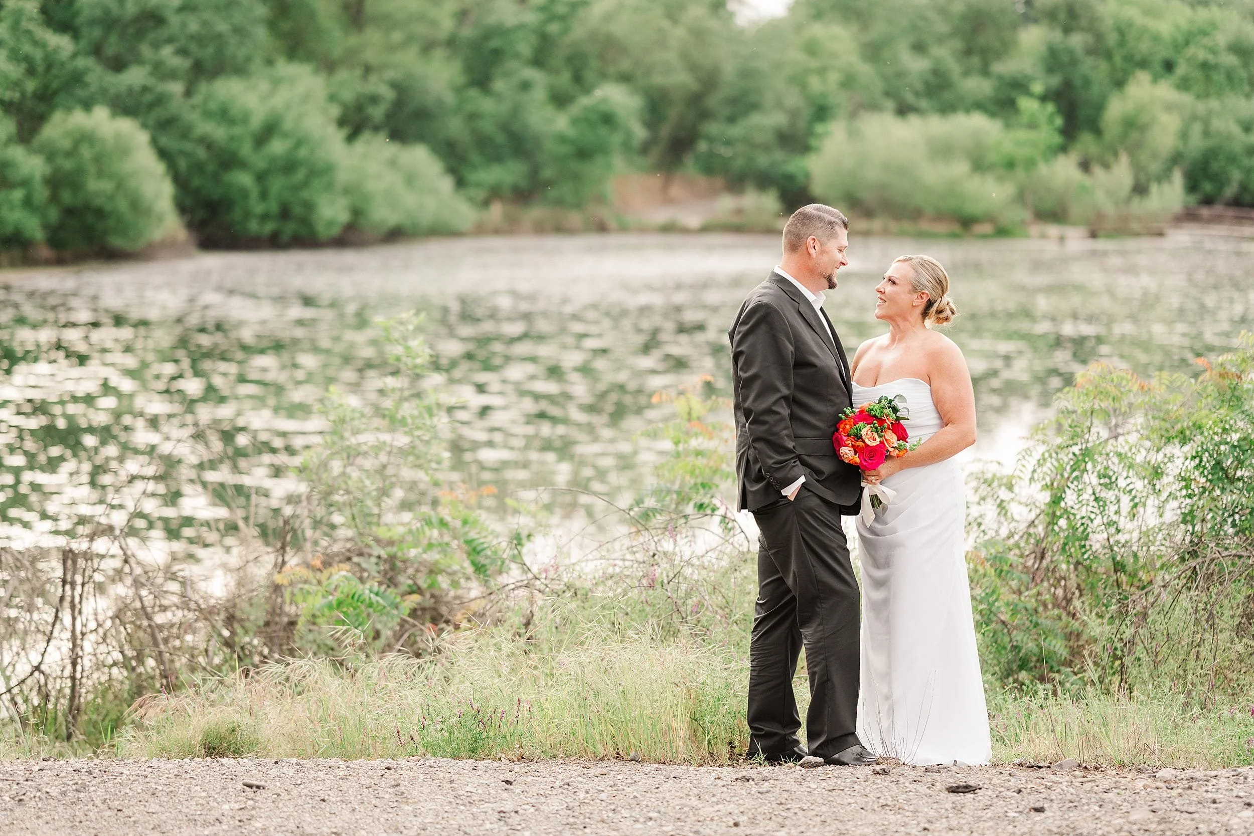 Bride in white dress holds red bouquet, groom in black suit by a serene lake with lush greenery; they gaze at each other, smiling warmly.