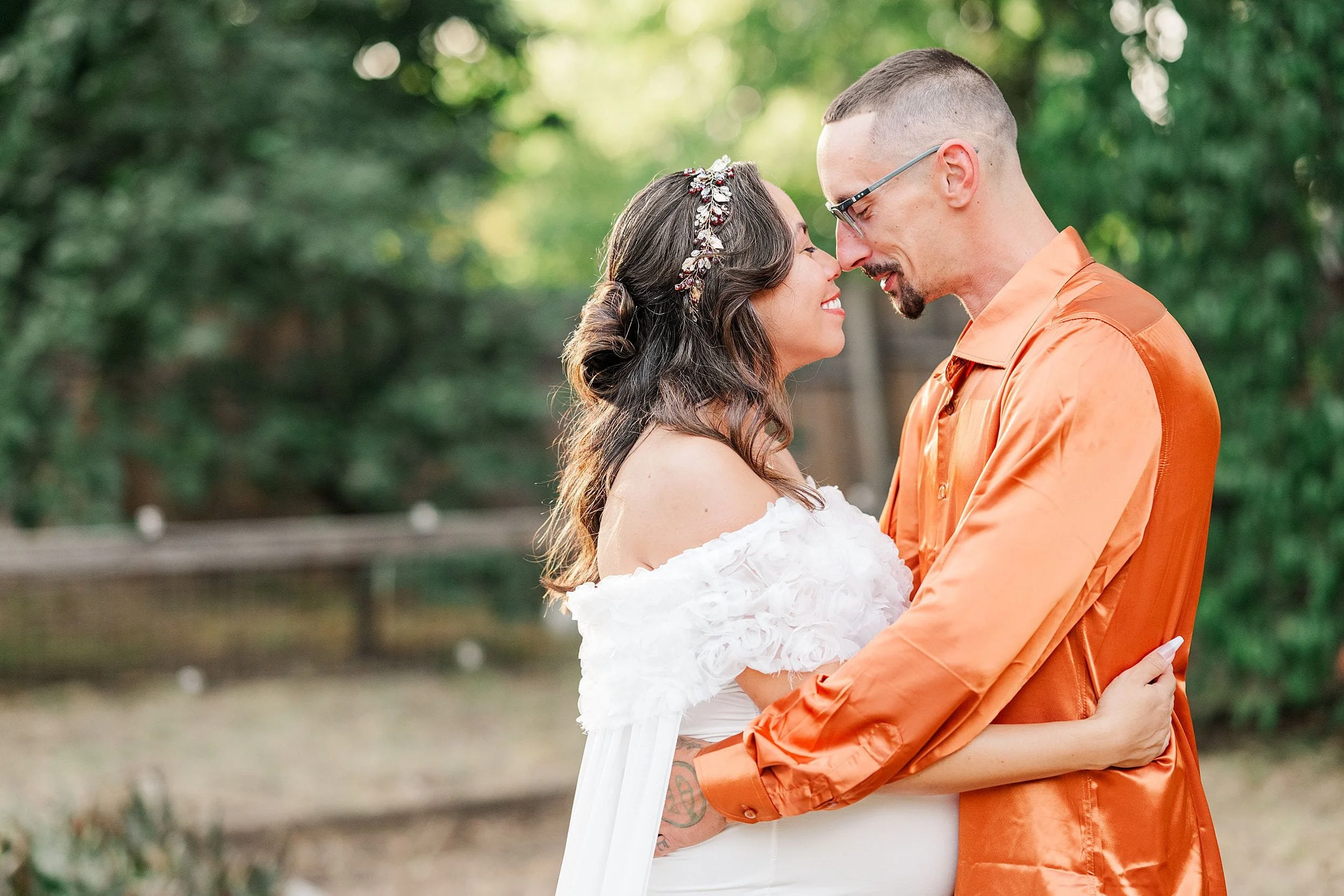 Newlywed couple embraces outdoors: bride in white dress and floral hairpiece gazes at groom in orange shirt, smiling in a sunlit forest.