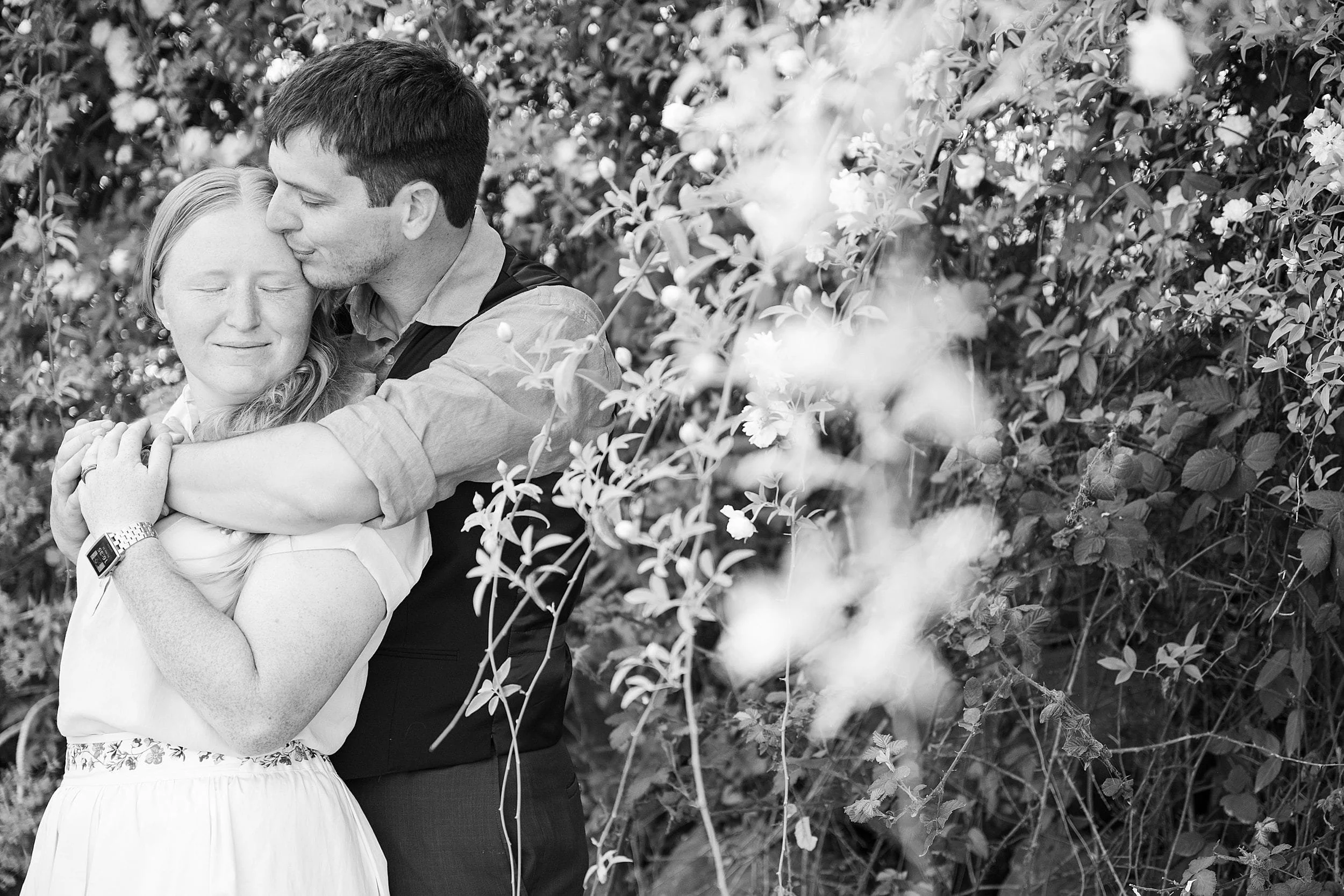 A couple embraces lovingly in a garden, surrounded by blooming flowers. The man gently kisses the woman's forehead, conveying warmth and affection.