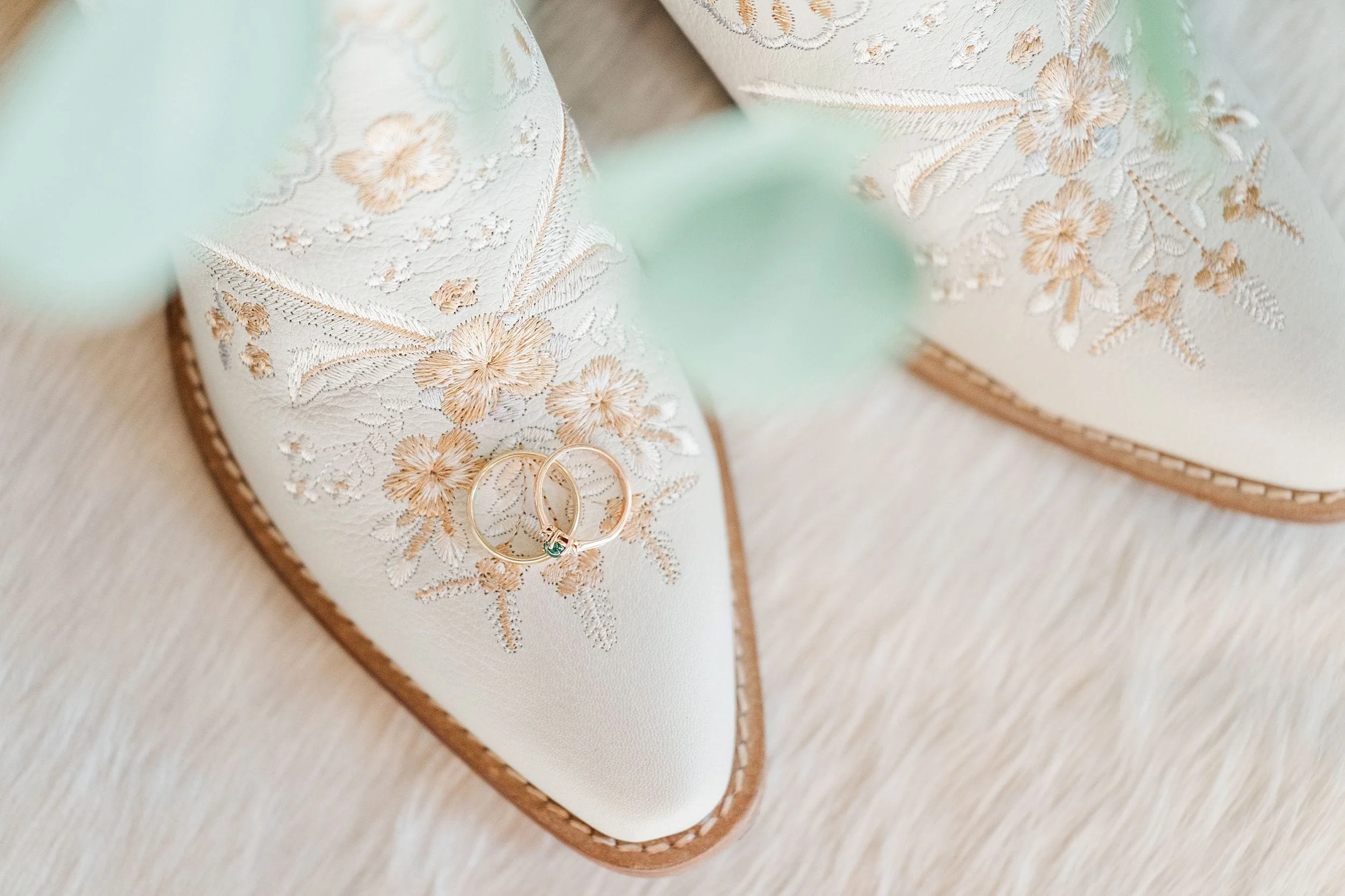 Ivory pointed-toe bridal boots with delicate gold floral embroidery and wedding rings resting on one toe, styled on soft fur for a romantic, elegant mood.