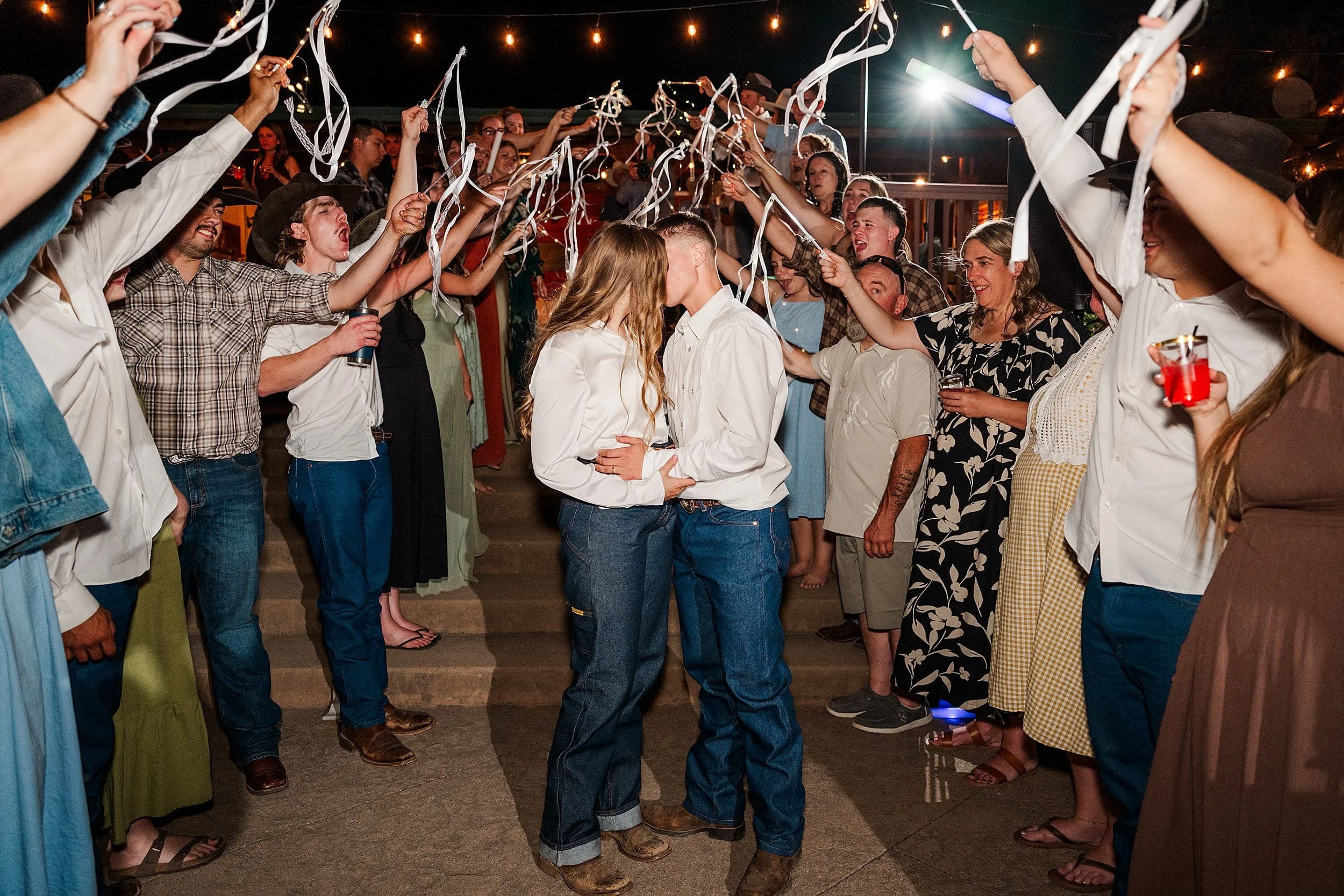 A couple kisses at an outdoor wedding party as guests wave silver streamers beneath glowing string lights, creating a joyful, festive mood.