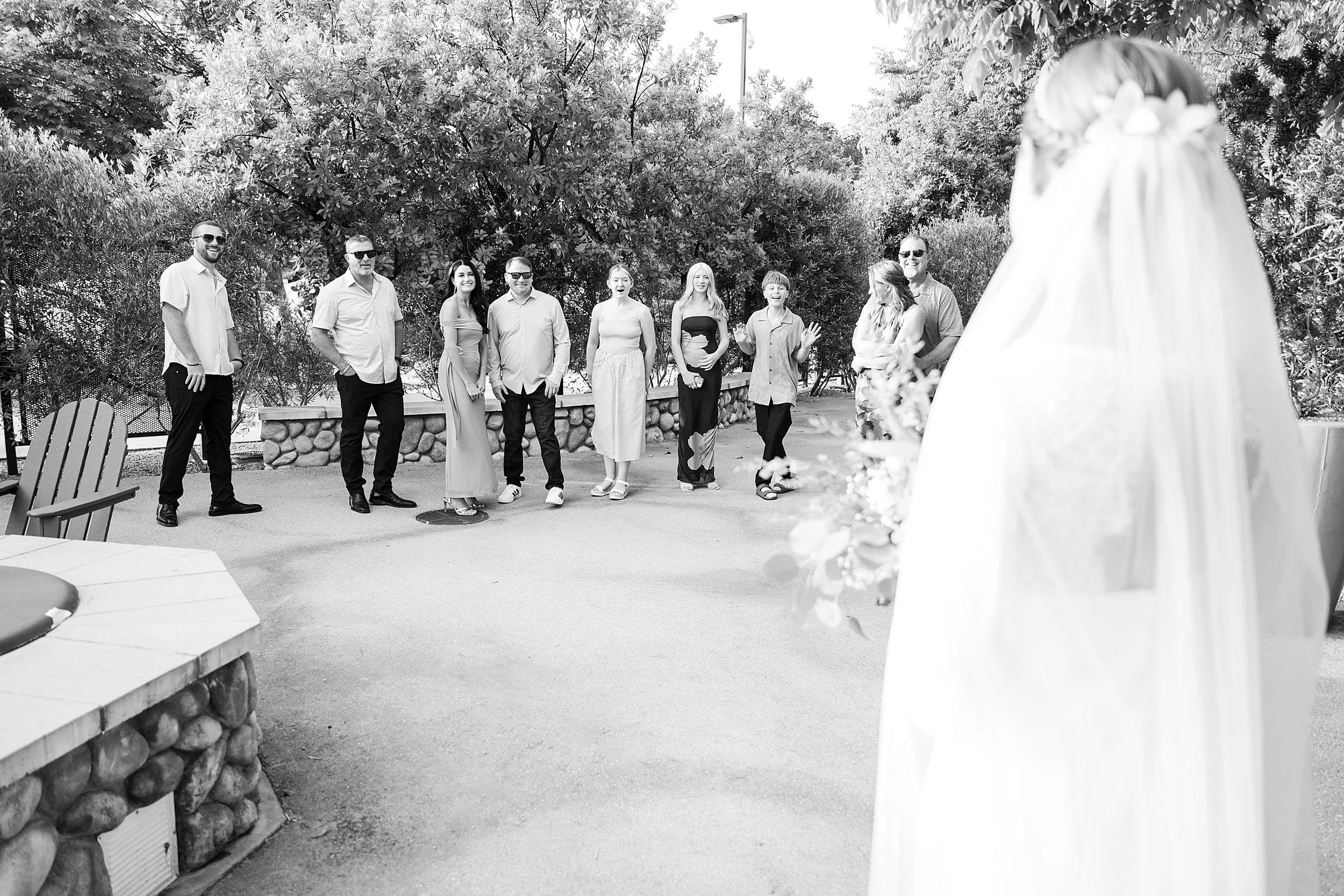 A bride, seen from behind with a flowing veil, approaches a group of smiling people outdoors. They stand together on a path surrounded by trees, creating a joyful, celebratory atmosphere.