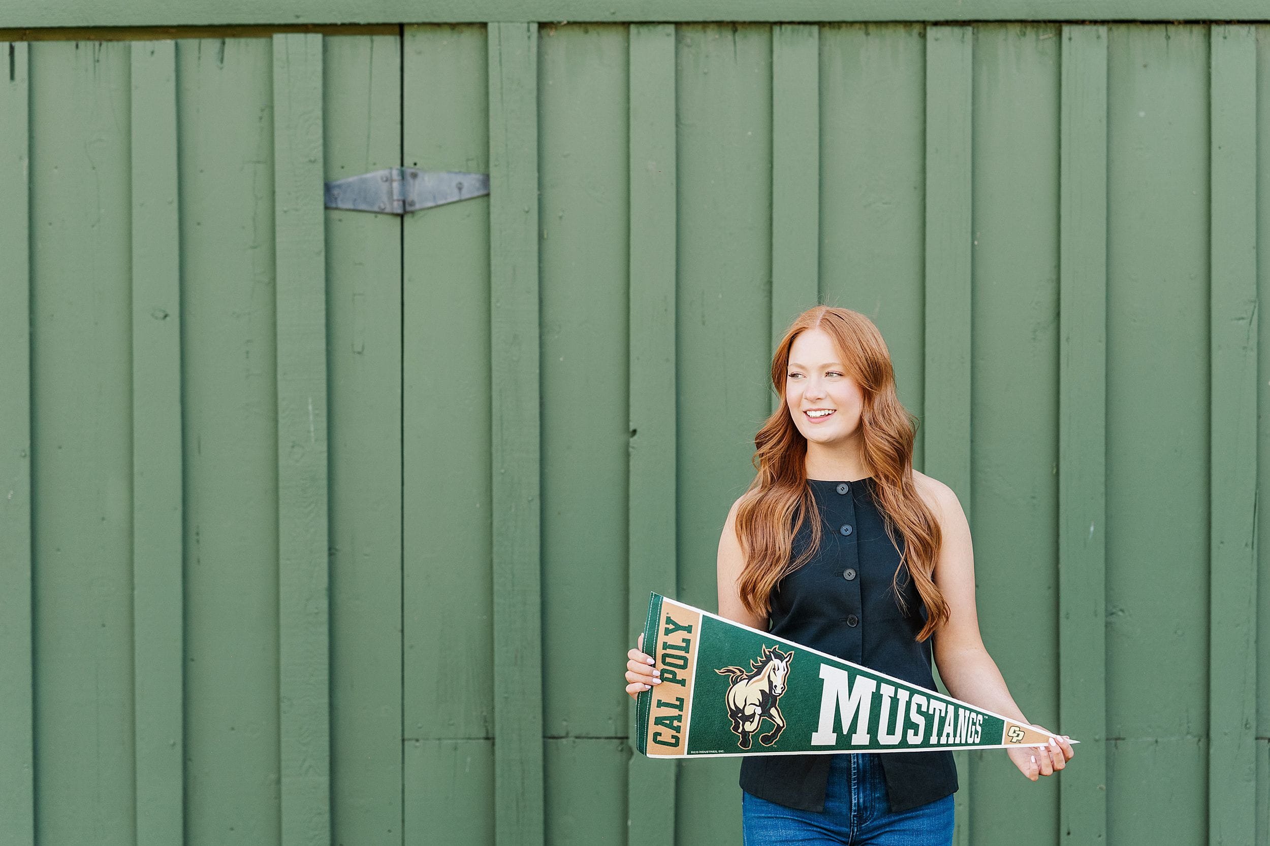 A person with long red hair smiles while holding a "Cal Poly Mustangs" pennant. The background features a green wooden wall, creating a cheerful tone.