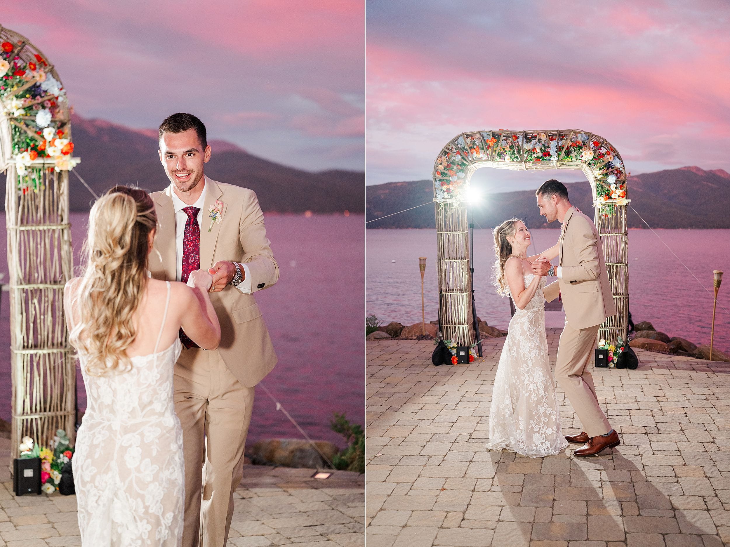 Bride and groom share a romantic moment beneath a floral wedding arch beside a lake at sunset, with pink clouds and mountains behind them.