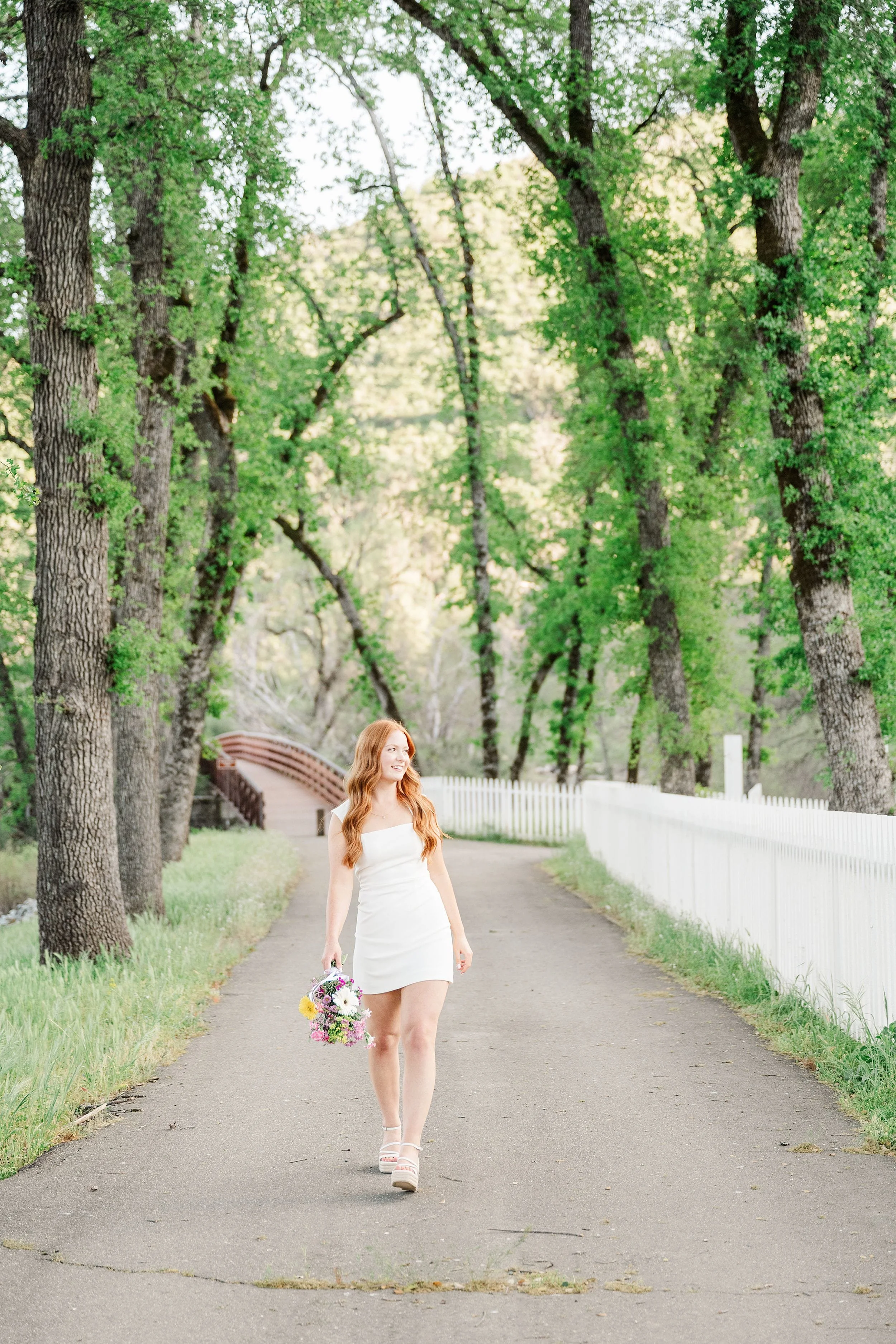 A woman in a white dress walks down a tree-lined path holding a bouquet. The scene is serene and picturesque, with vibrant green foliage and a white fence.