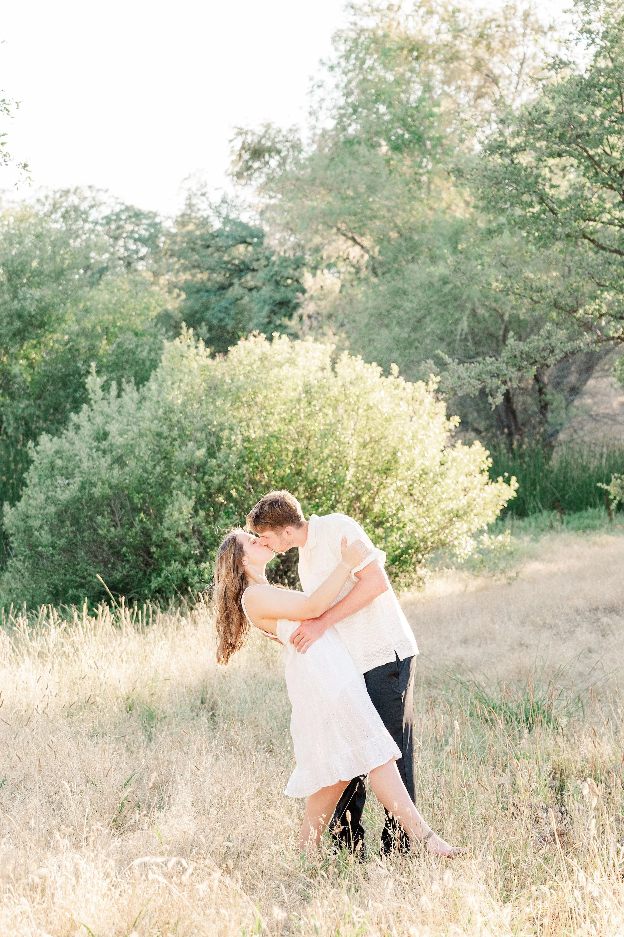 A couple shares a romantic kiss in a sunlit field surrounded by lush green trees. The woman wears a white dress; the man in light clothing dips her gently.