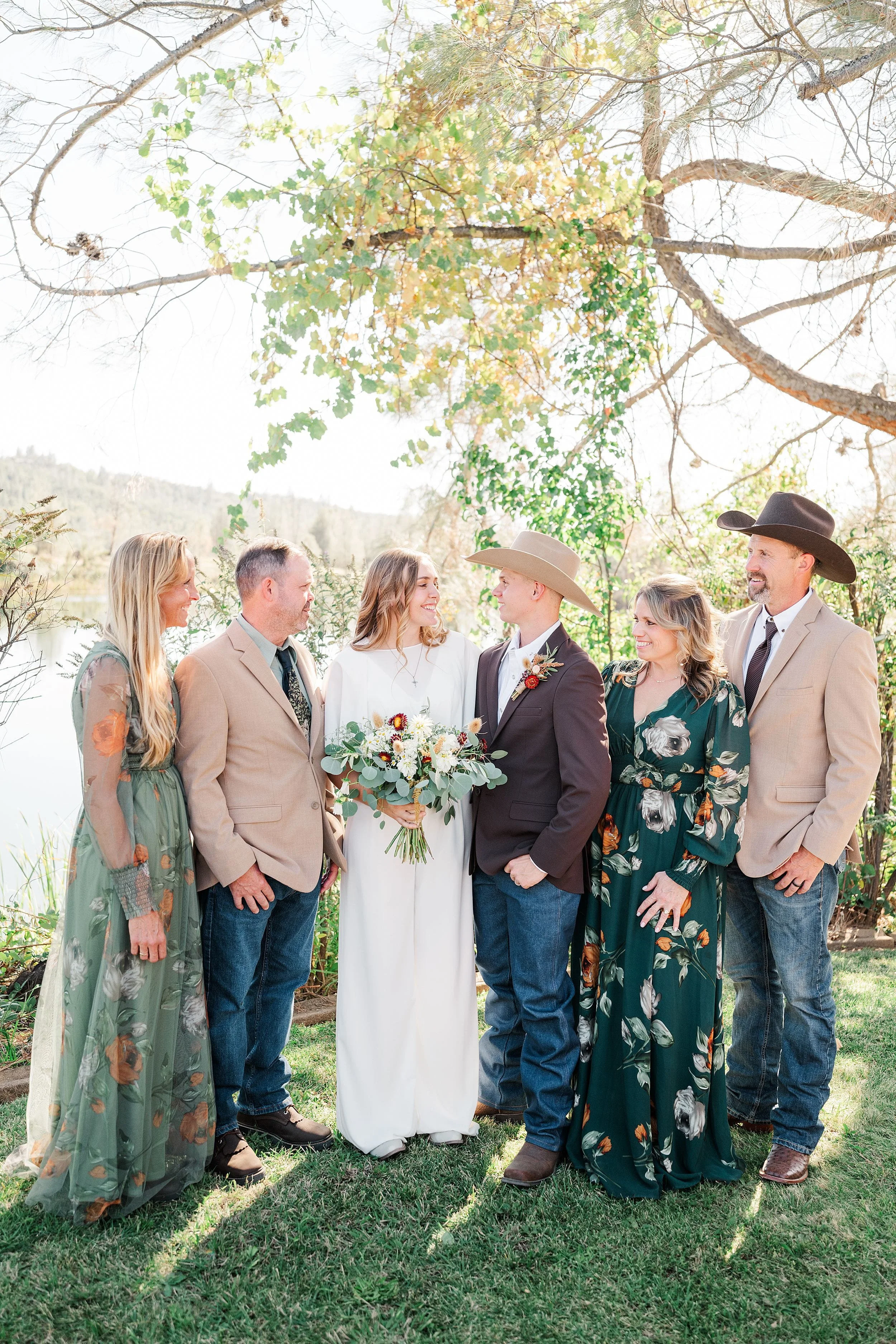 A bride and groom stand with their parents beneath a leafy tree at an outdoor wedding, sharing warm smiles in soft natural light.
