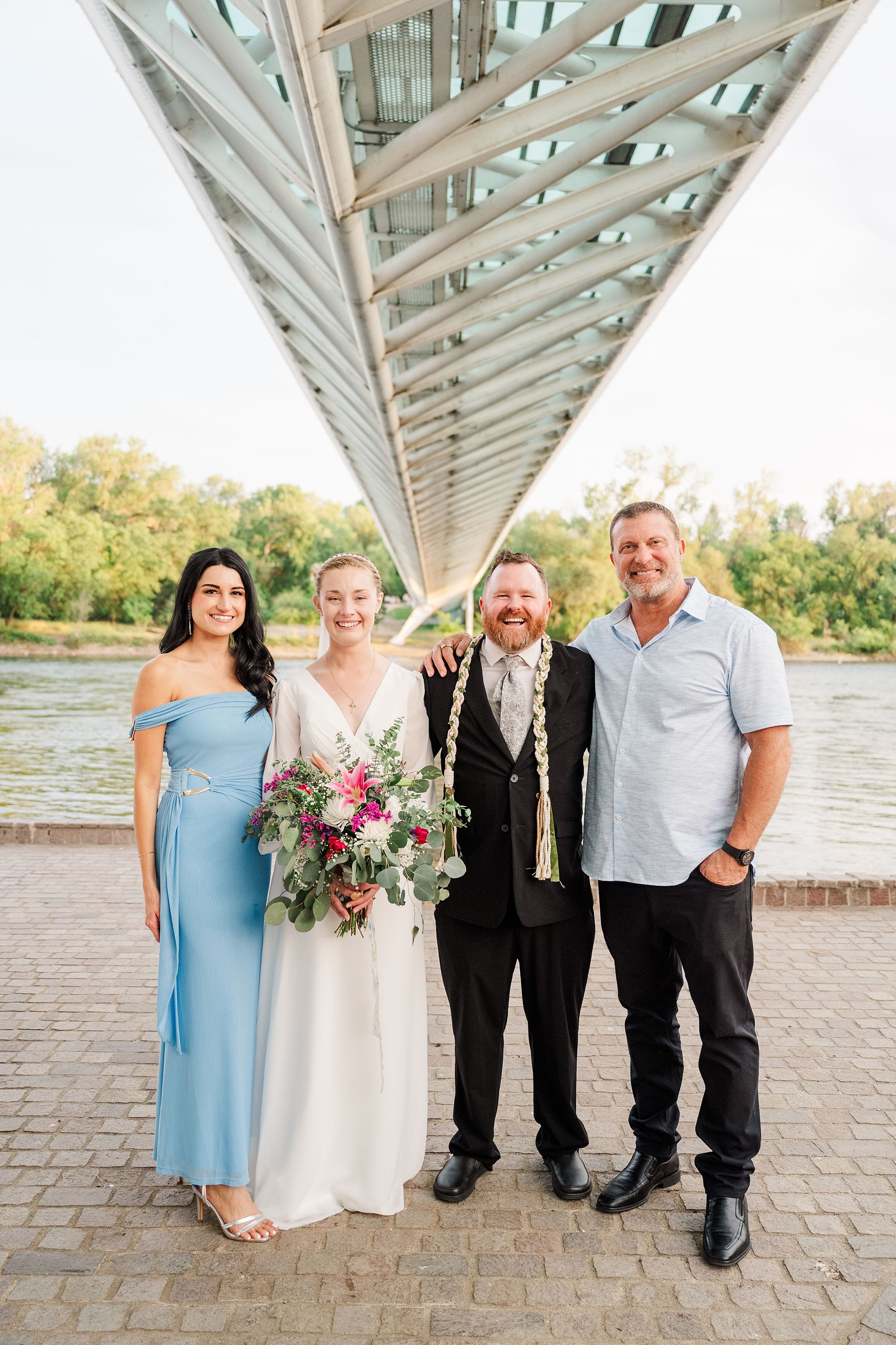 Four people stand under a modern bridge beside a river. A woman in a white dress holds a vibrant bouquet, and everyone smiles warmly, conveying joy and celebration.