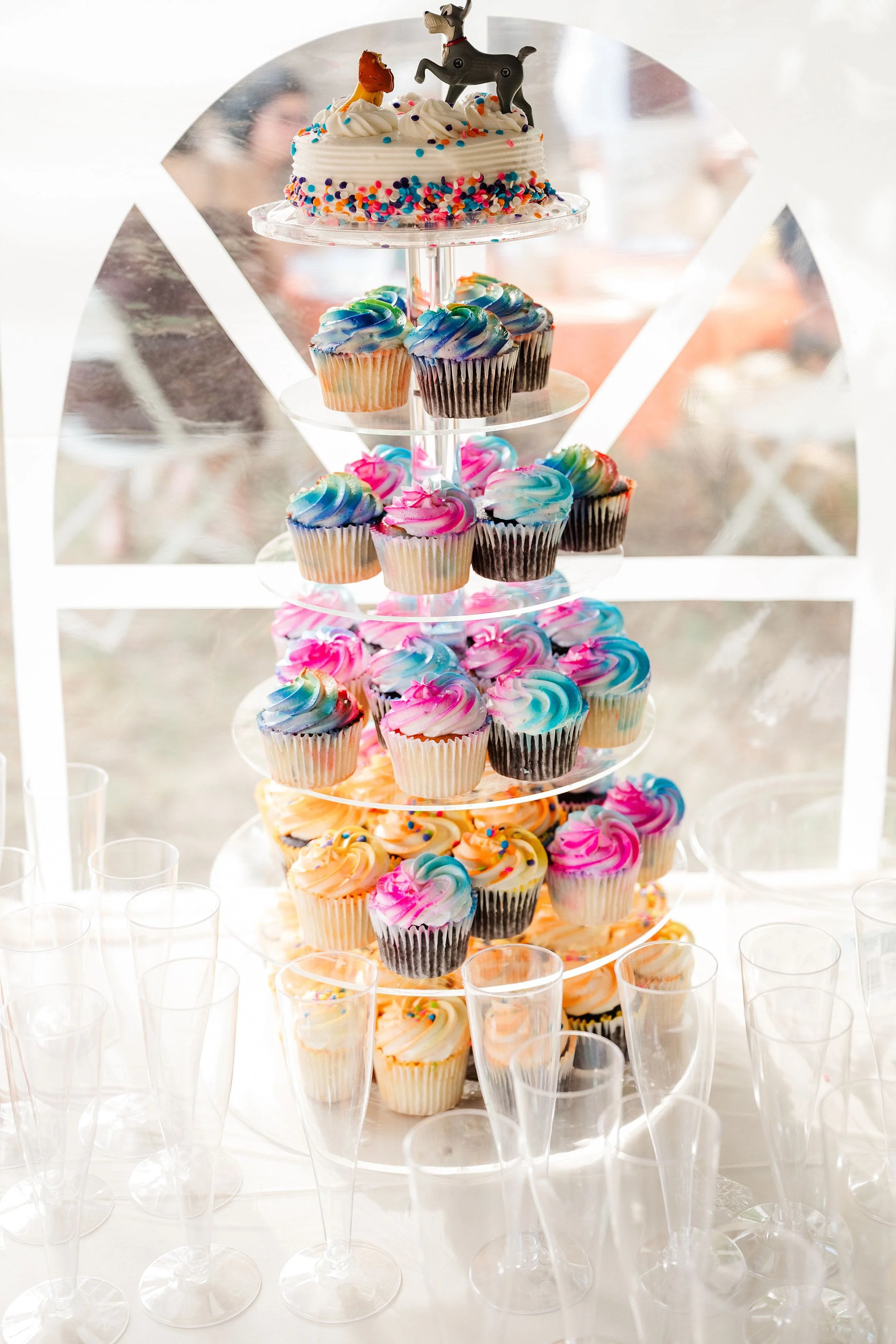 Festive tiered dessert stand with a decorated cake and colorful rainbow-frosted cupcakes, surrounded by empty glasses at a bright, cheerful party.