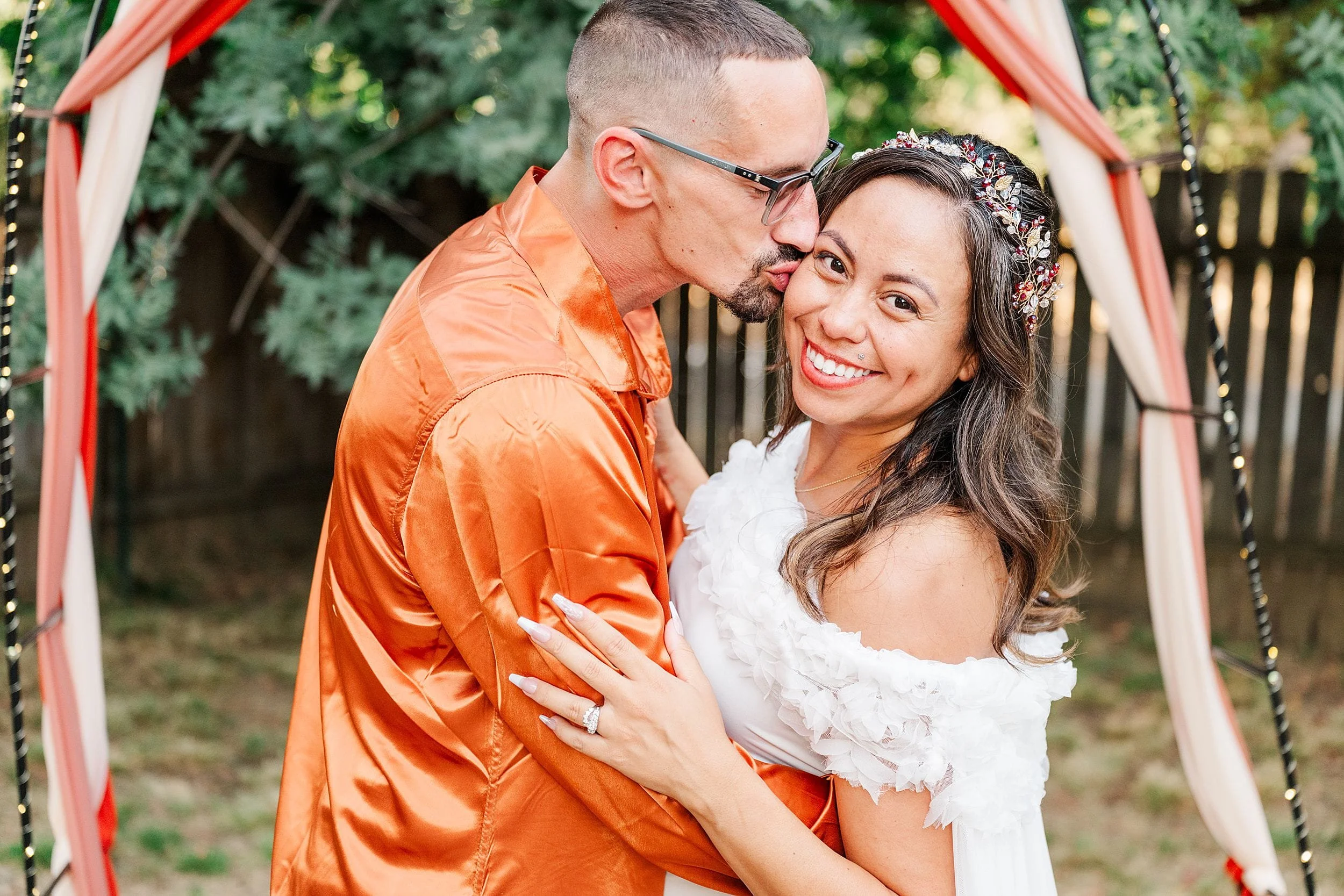 Newlywed couple embrace beneath a draped wedding arch; groom kisses the bride’s cheek as she smiles in a white dress and floral crown.