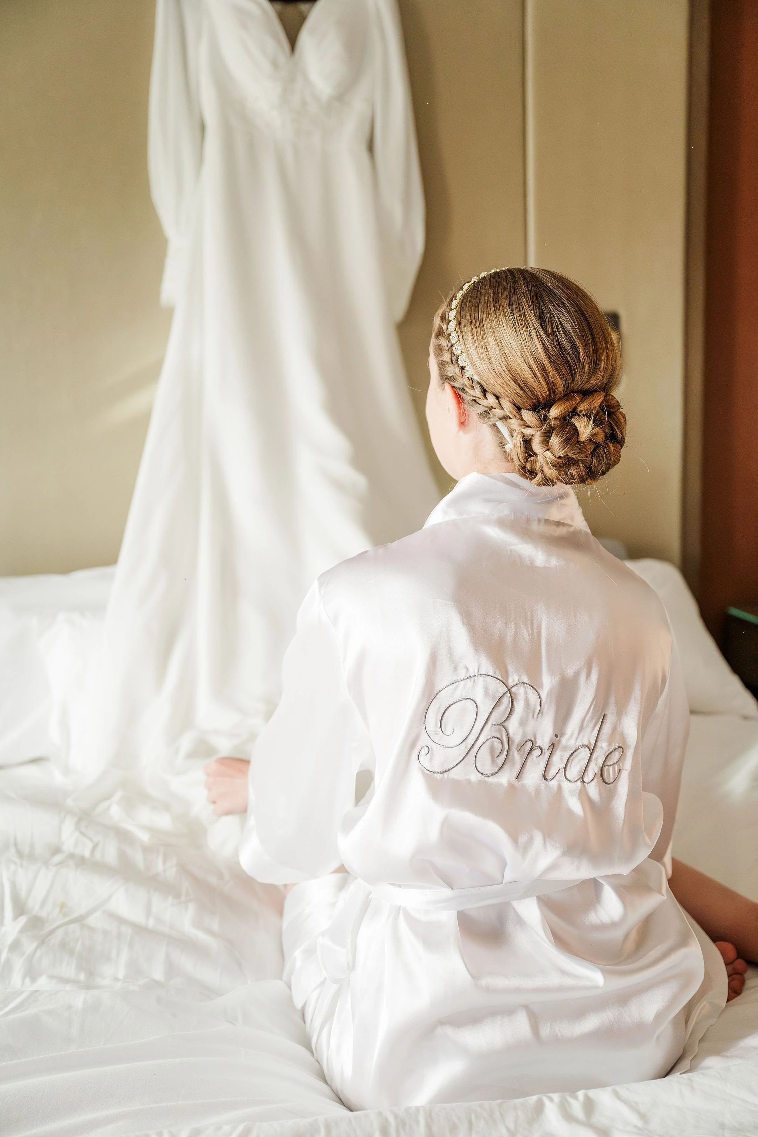 A bride in a white robe sits on a bed, facing her wedding dress hanging nearby. Her hair is in a braided updo, creating a serene and anticipatory mood.
