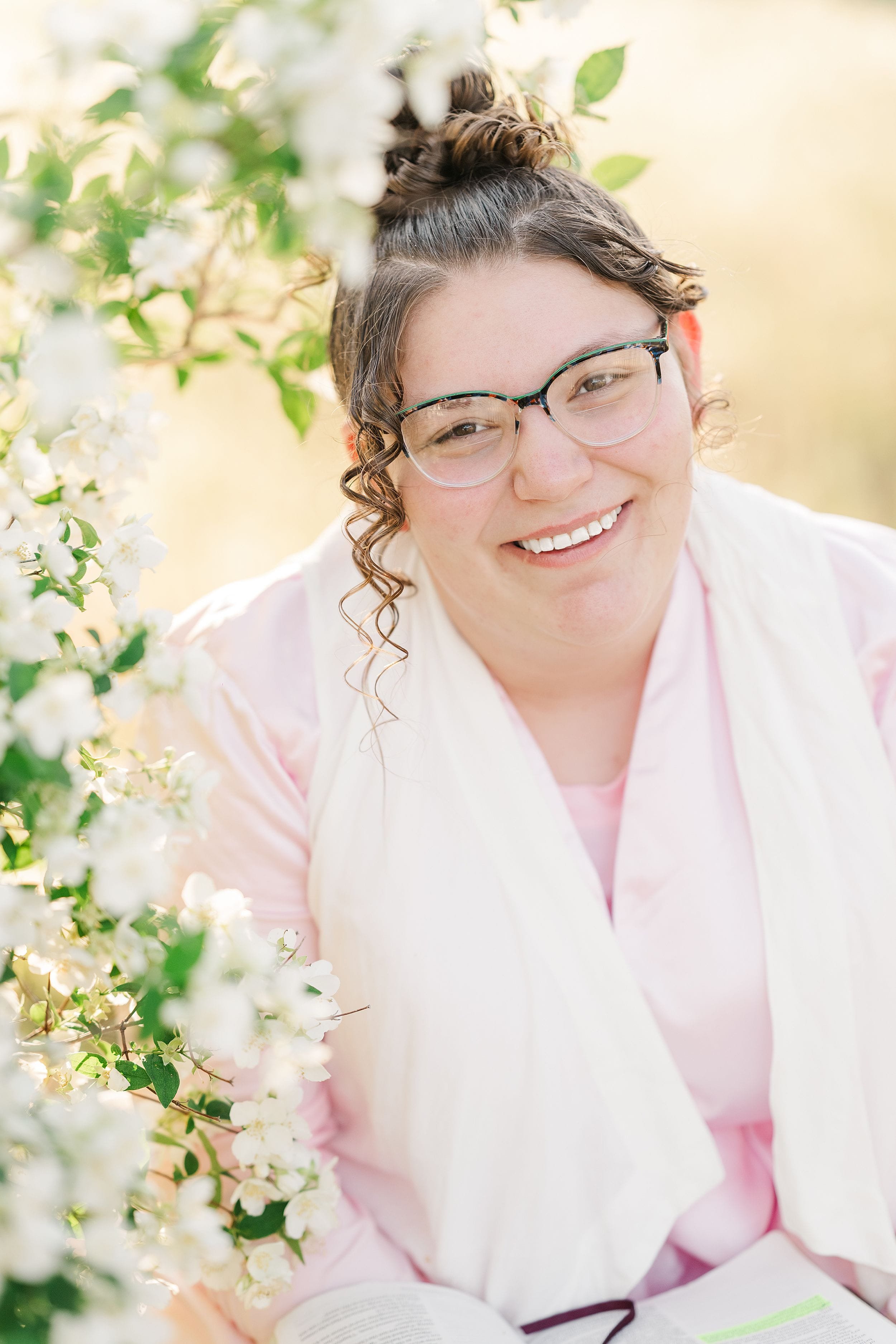 A smiling person with glasses and curly hair sits outdoors, surrounded by blossoming white flowers. They wear a light pink shirt and white scarf, conveying a serene and joyful mood.