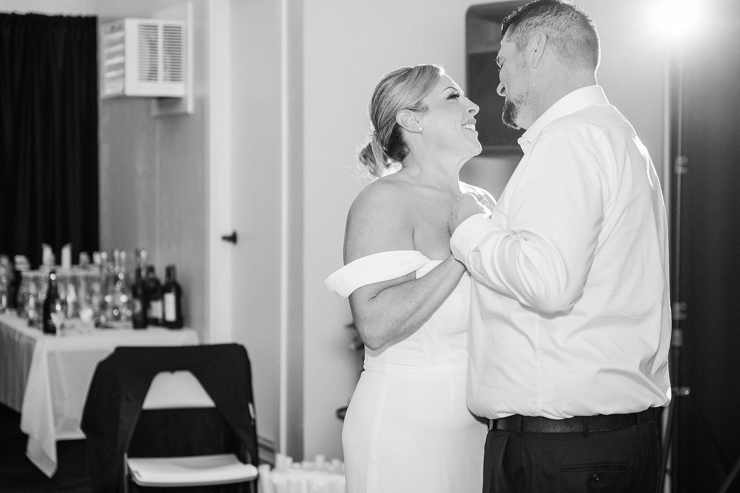 A smiling couple dances closely in a softly lit room, surrounded by tables with bottles. The atmosphere is joyful and intimate, with a monochrome effect.