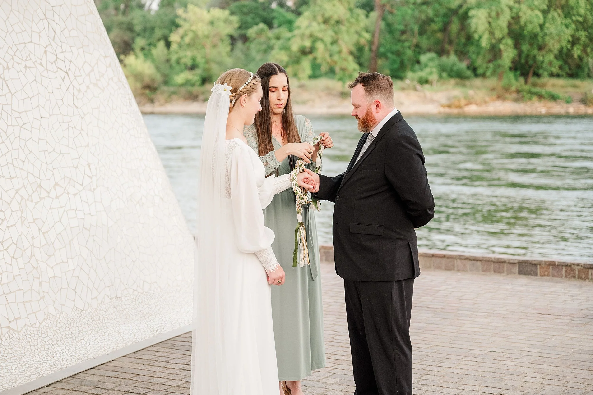 A bride and groom stand by a river, their hands tied with a ribbon by a woman in a green dress. The scene is serene and intimate, with lush greenery in the background.