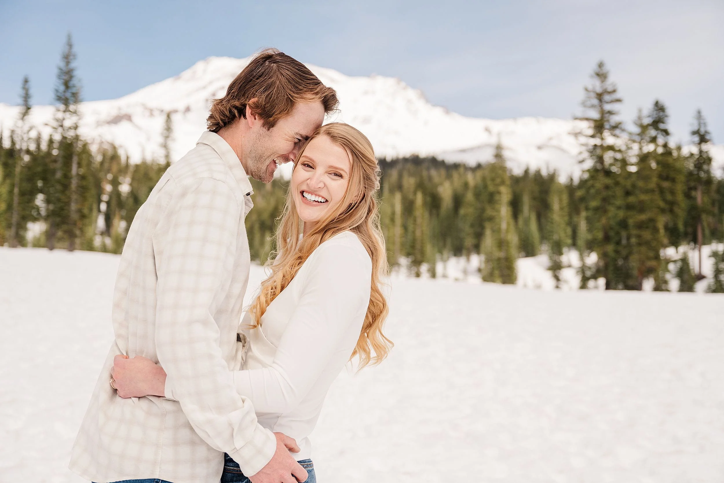 A couple embraces in a snowy landscape, with mountains and pine trees in the background. They appear joyful, smiling warmly under a clear blue sky.