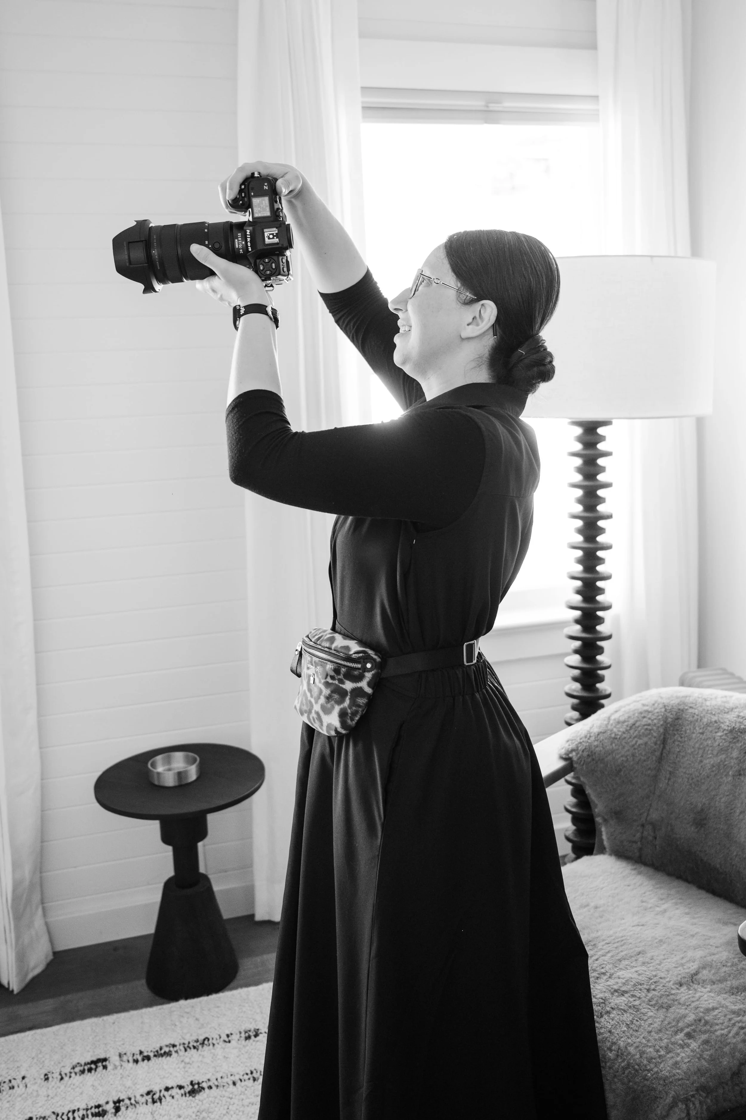 A woman with dark hair and glasses is smiling while holding a camera in her hand. She is wearing a black long-sleeve top and a skirt with a small leopard print bag at her waist. The room has white walls, curtains, a bed, a side table with a round metallic object, a patterned rug, and a lamp with a spiral base.