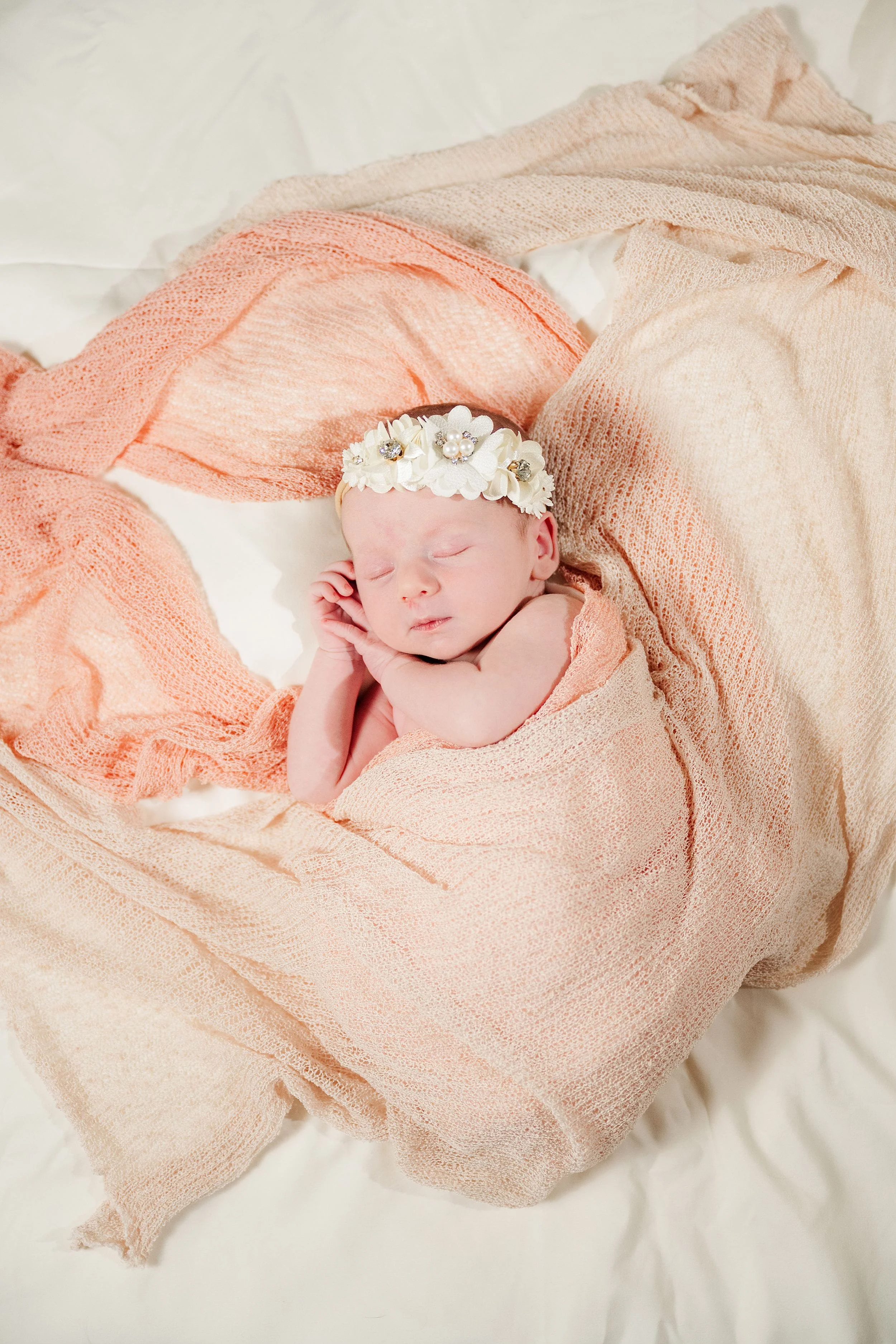 Newborn baby peacefully sleeping, wrapped in soft, peach-colored fabric with a floral headband, conveying warmth and serenity.