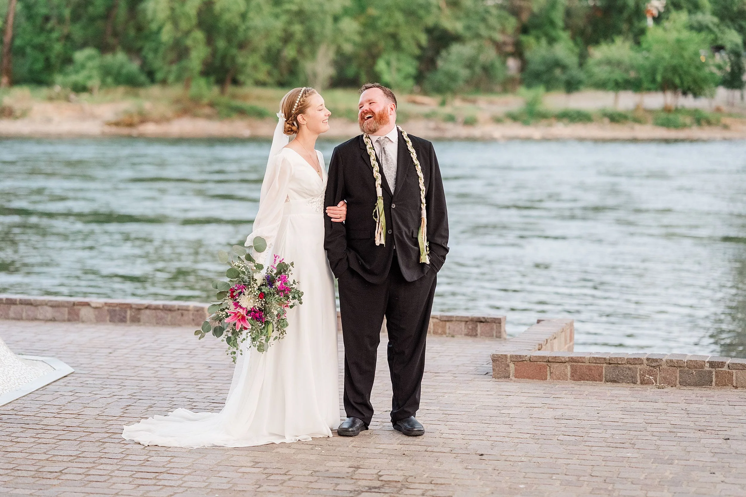 A joyful bride in a flowing white gown holds a vibrant bouquet, linking arms with a groom in a black suit by a serene riverside with lush greenery.