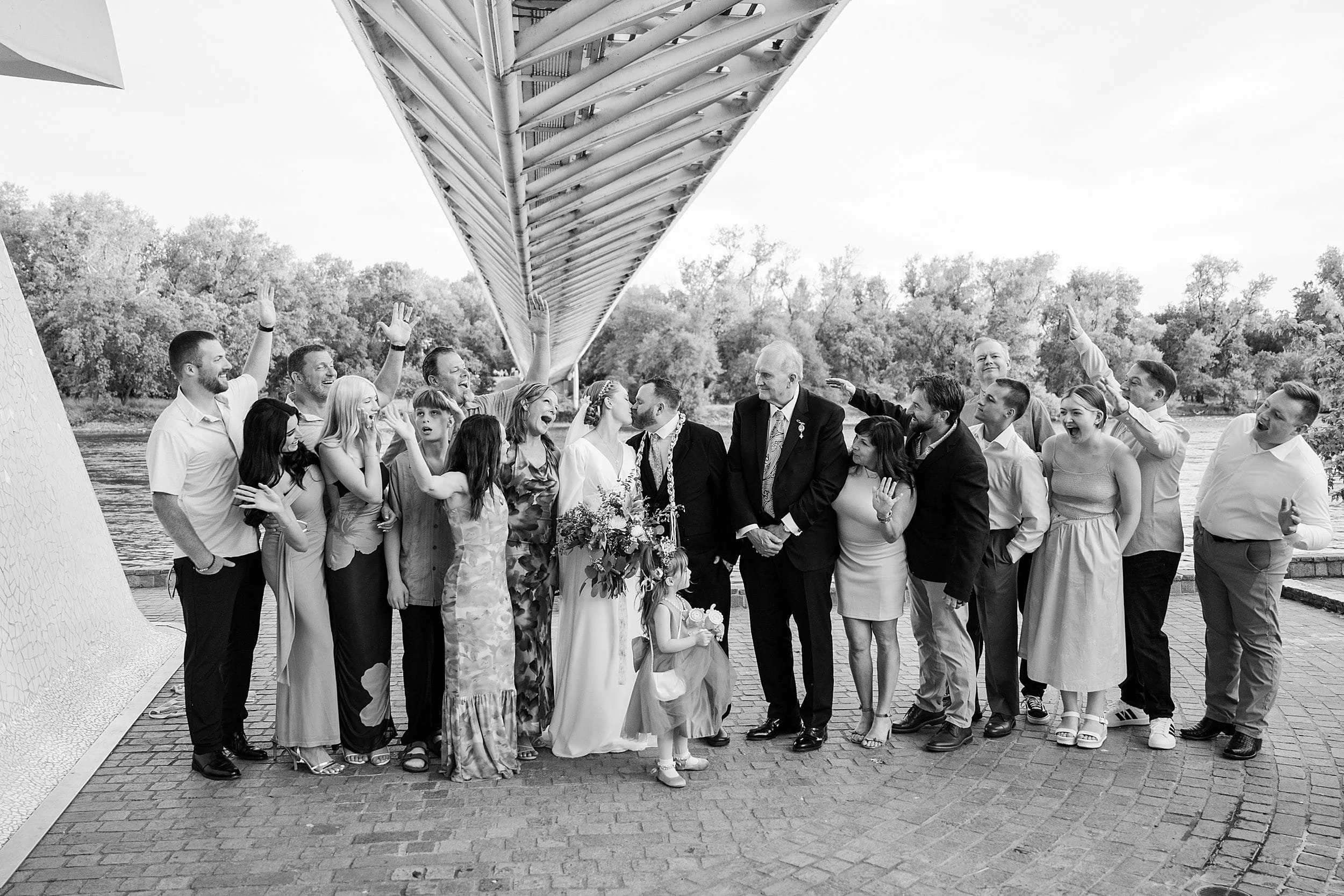 A joyful black-and-white wedding scene shows a smiling group under a geometric bridge. Guests, in celebratory poses, surround the bride and groom.