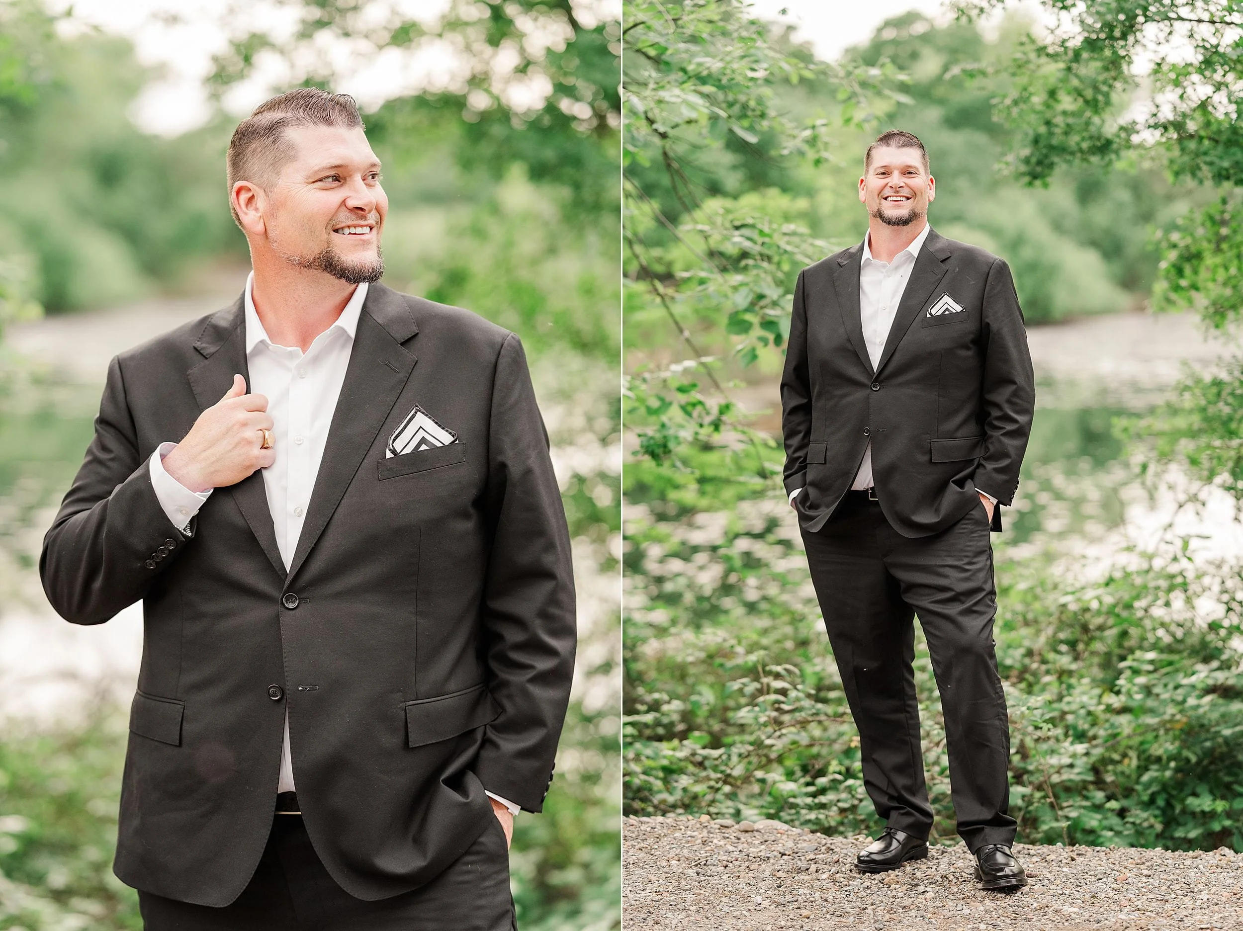 Man in a black suit with a white shirt and pocket square stands outdoors. He smiles confidently, surrounded by lush greenery and a calm river.