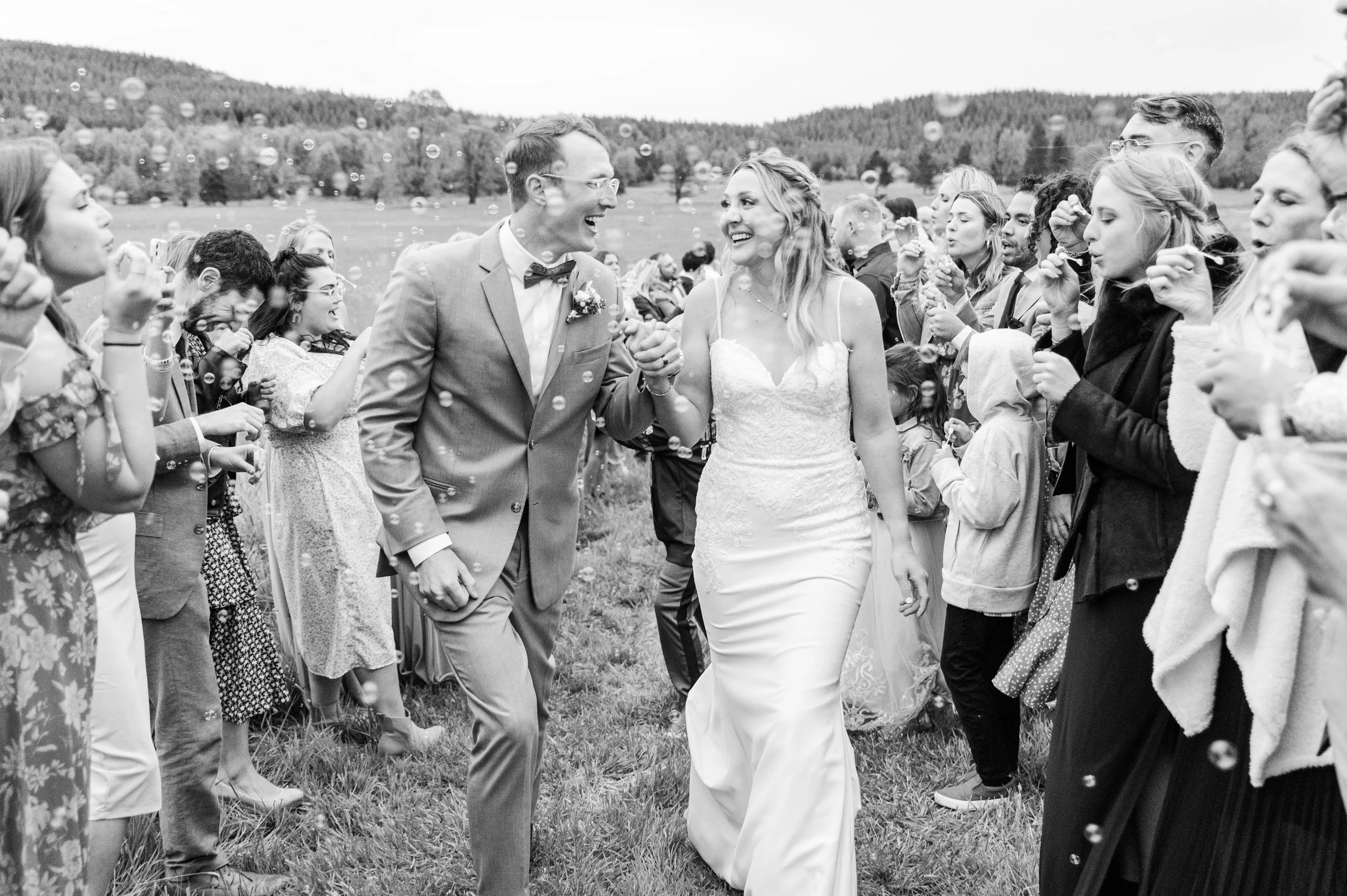 A bride and groom walking hand in hand through a crowd of wedding guests, smiling, during their outdoor wedding celebration, with mountains in the background.