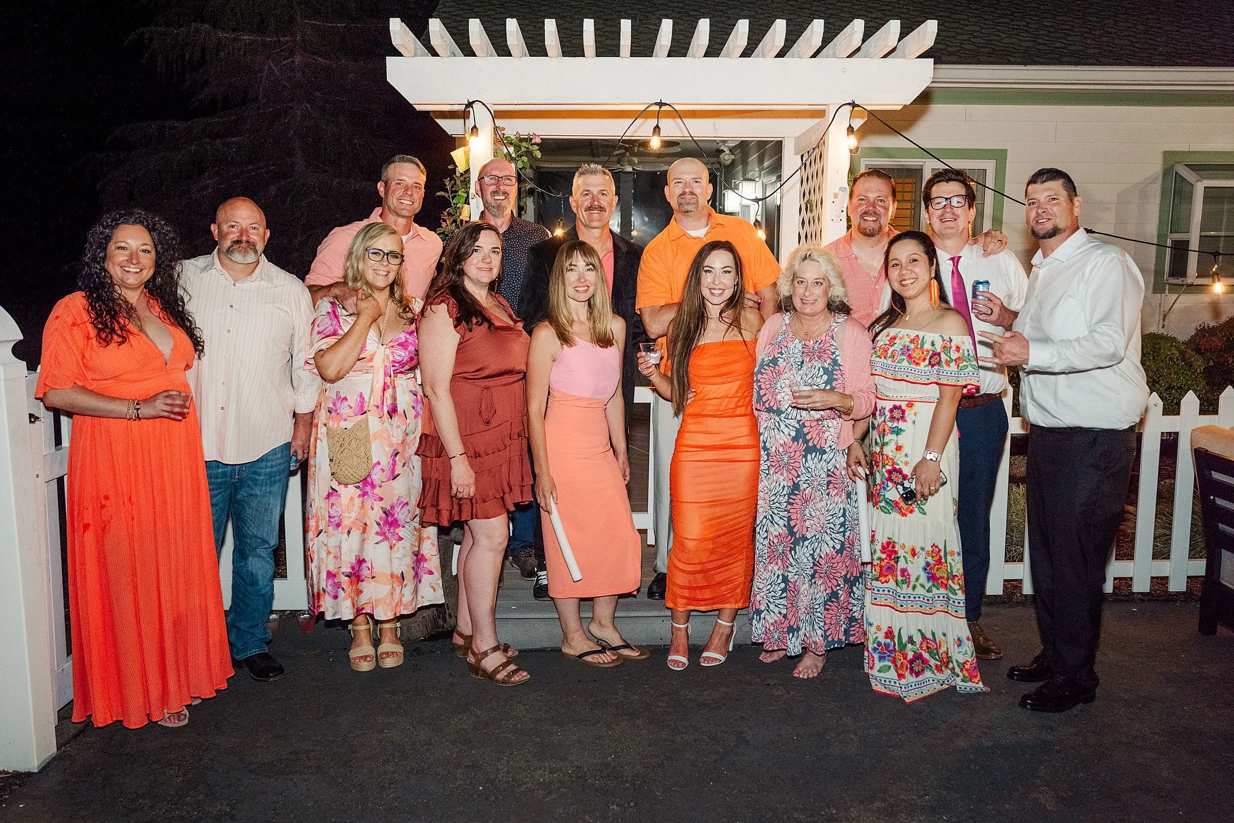 A group of 15 people stand together, smiling in front of a white pergola at night. Many wear bright or floral outfits, conveying a festive mood.