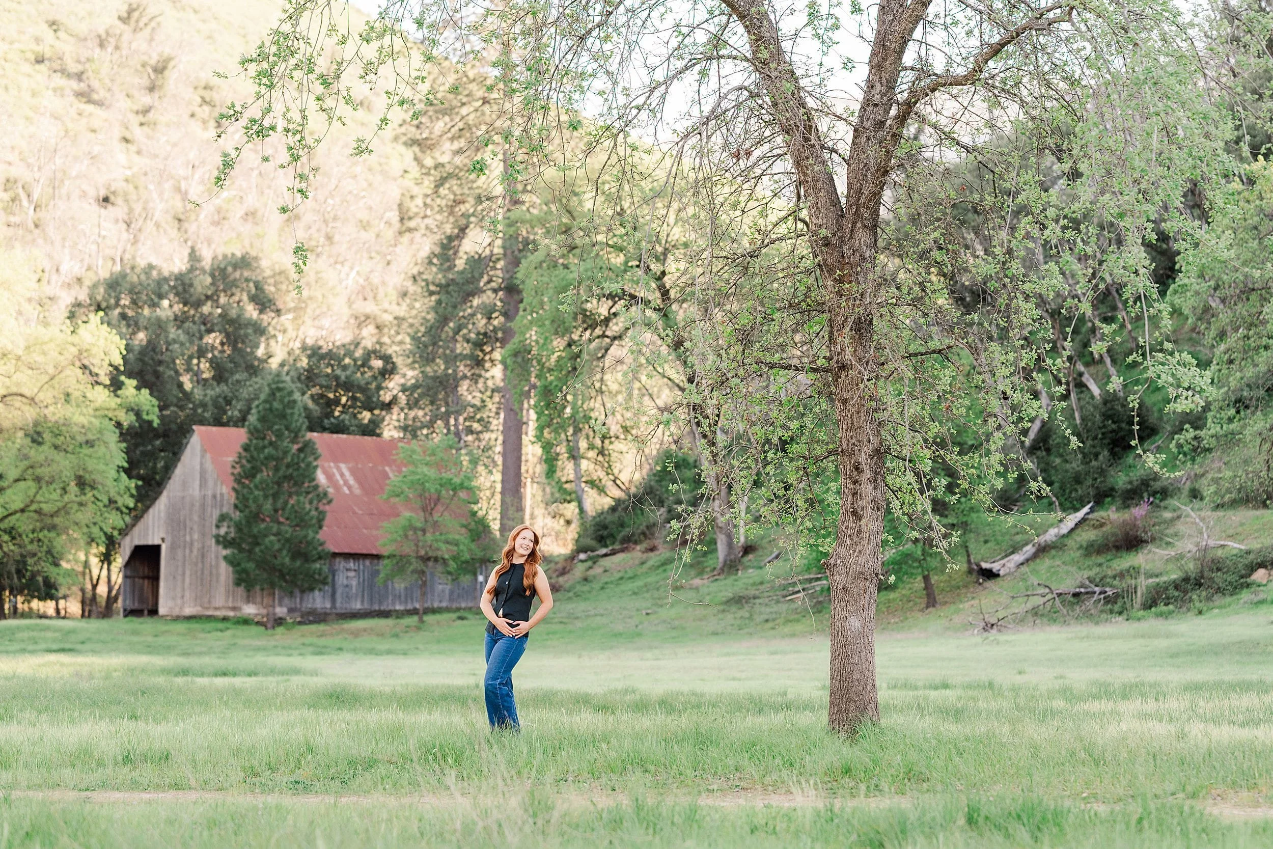A woman stands in a green field near a large tree, with a rustic barn and forested hillside in the background, conveying a serene rural scene.