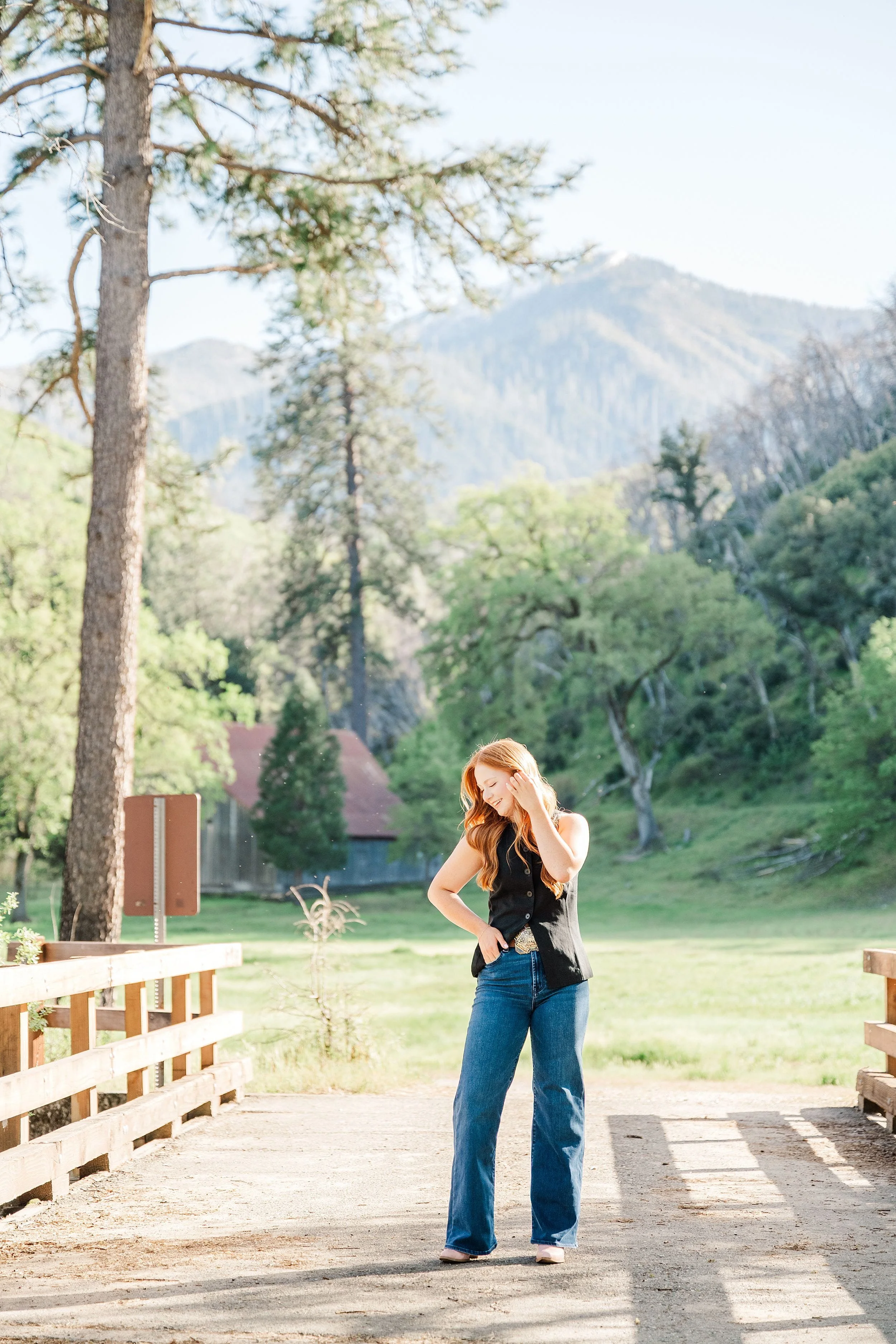 A woman with long hair stands on a sunlit wooden bridge in a forest setting, surrounded by lush greenery and distant mountains, exuding a peaceful vibe.