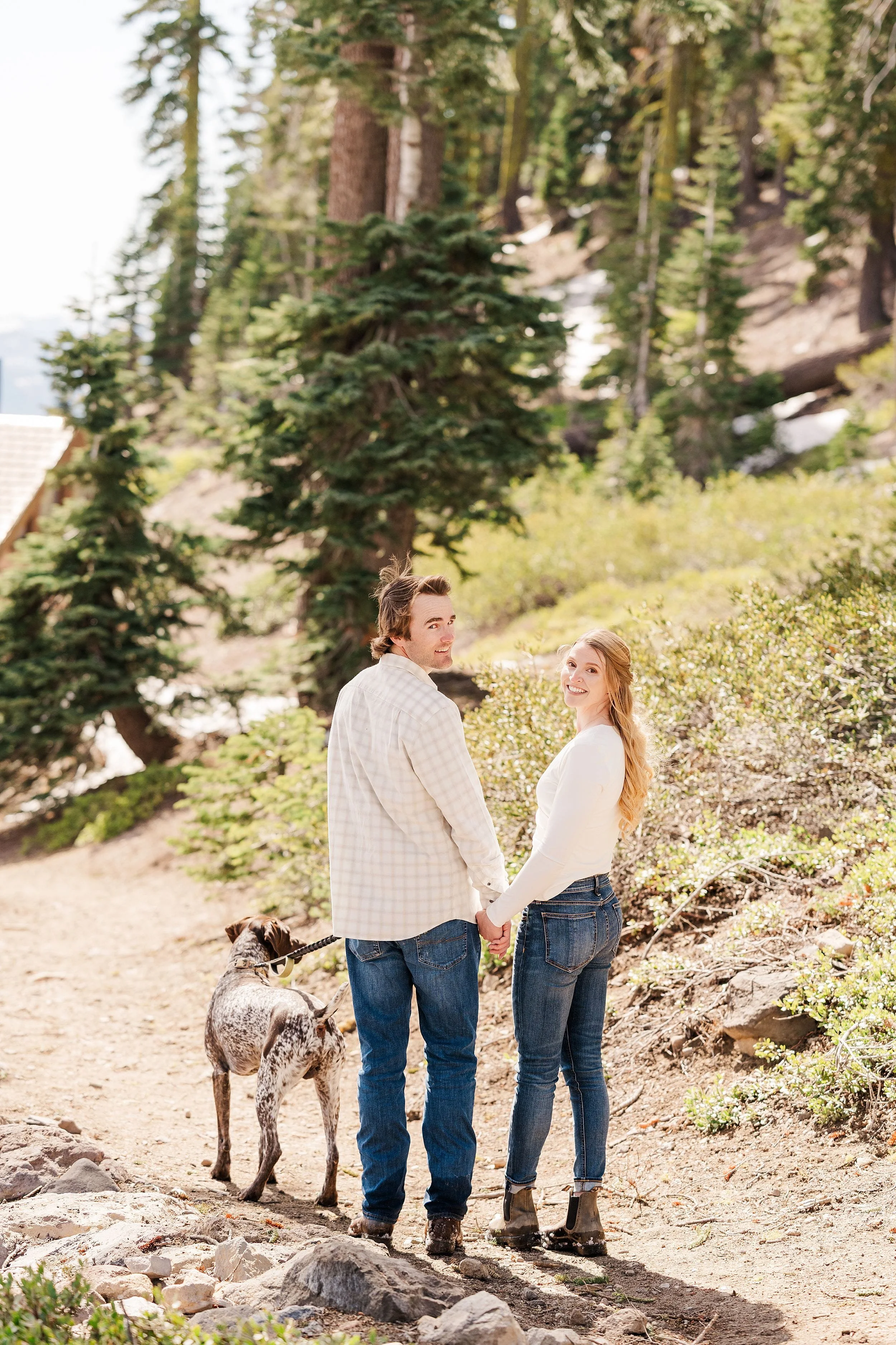 A couple holding hands and smiling back, walking a dog on a forest trail. They're surrounded by green trees and sunlight, conveying a joyful, serene mood.