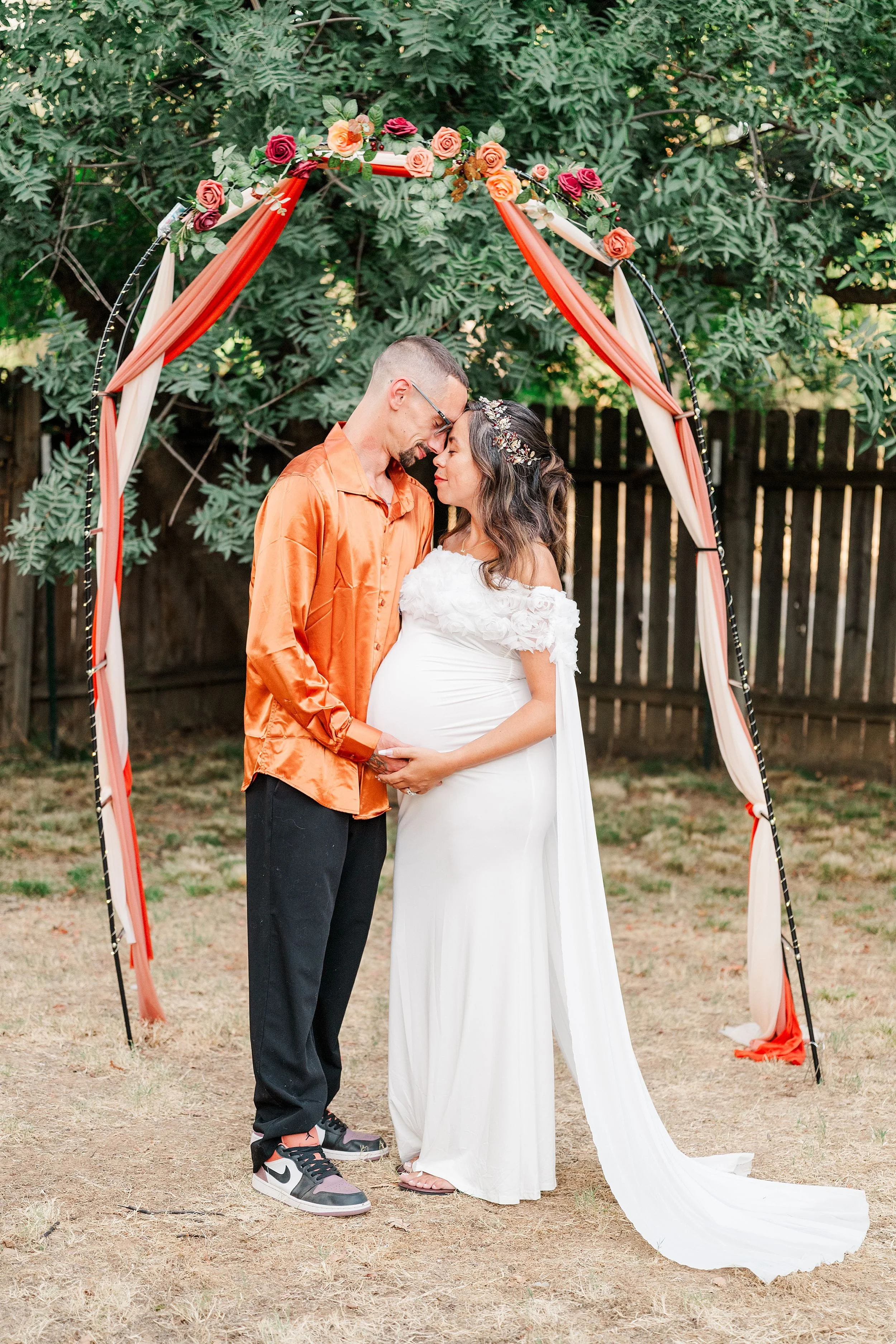 Pregnant couple kisses beneath a floral arch draped in red fabric outdoors, conveying tenderness and joy during a maternity celebration.