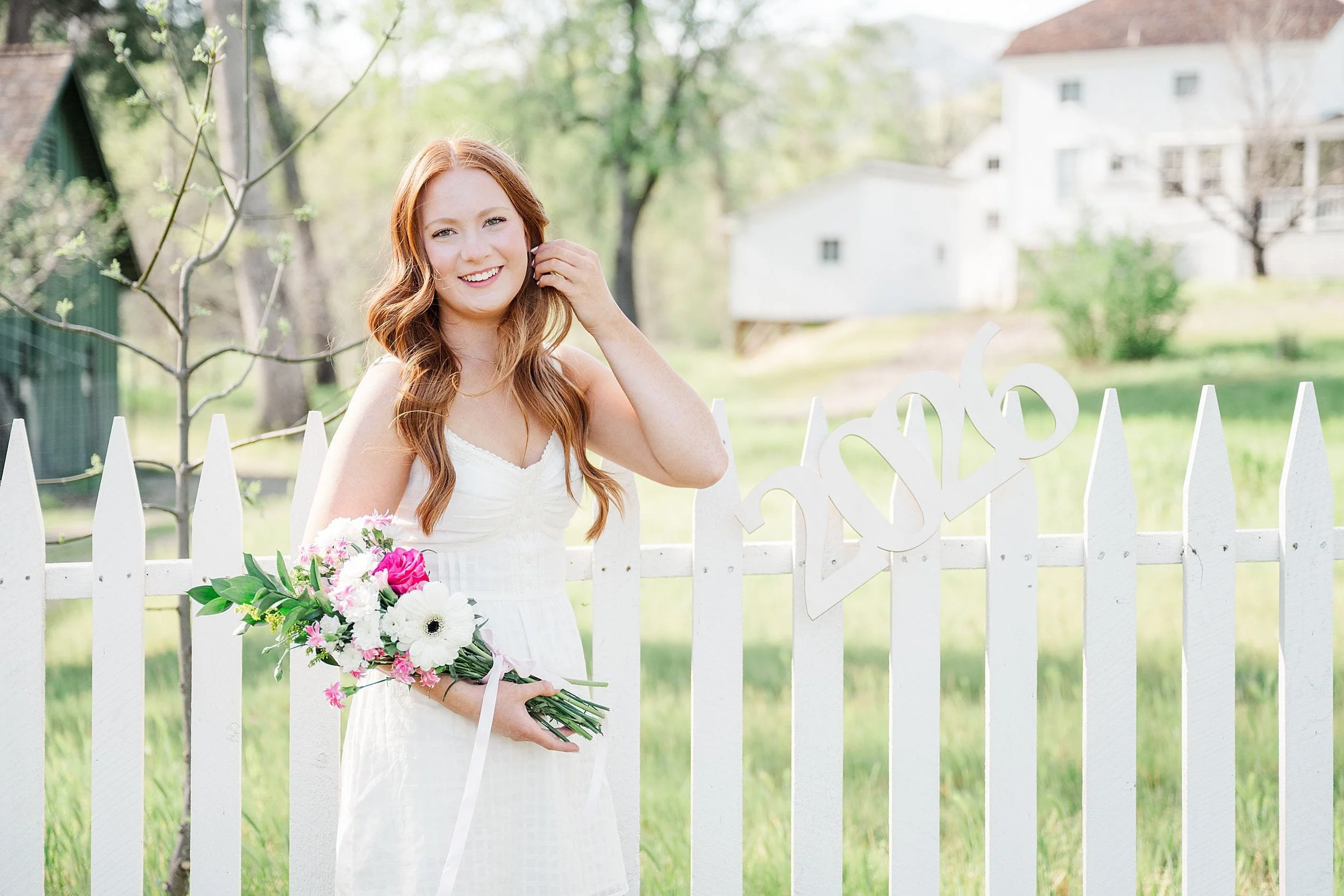 Young woman in a white dress smiling, holding a bouquet of flowers by a white picket fence with "2026" decoration. Background is a lush, sunny garden.