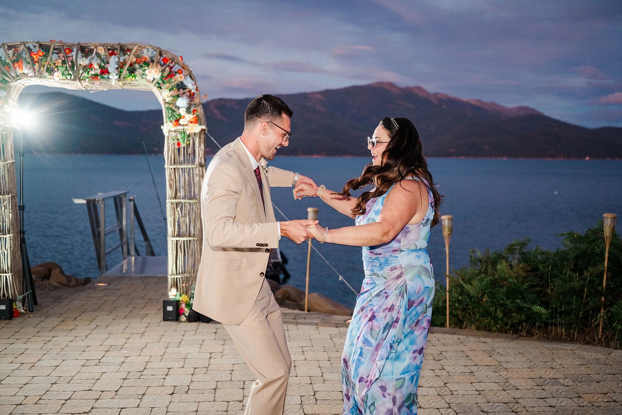 Mother and son dance beneath a flower-covered wedding arch beside a lake at twilight, with mountains behind them and a joyful, romantic mood.