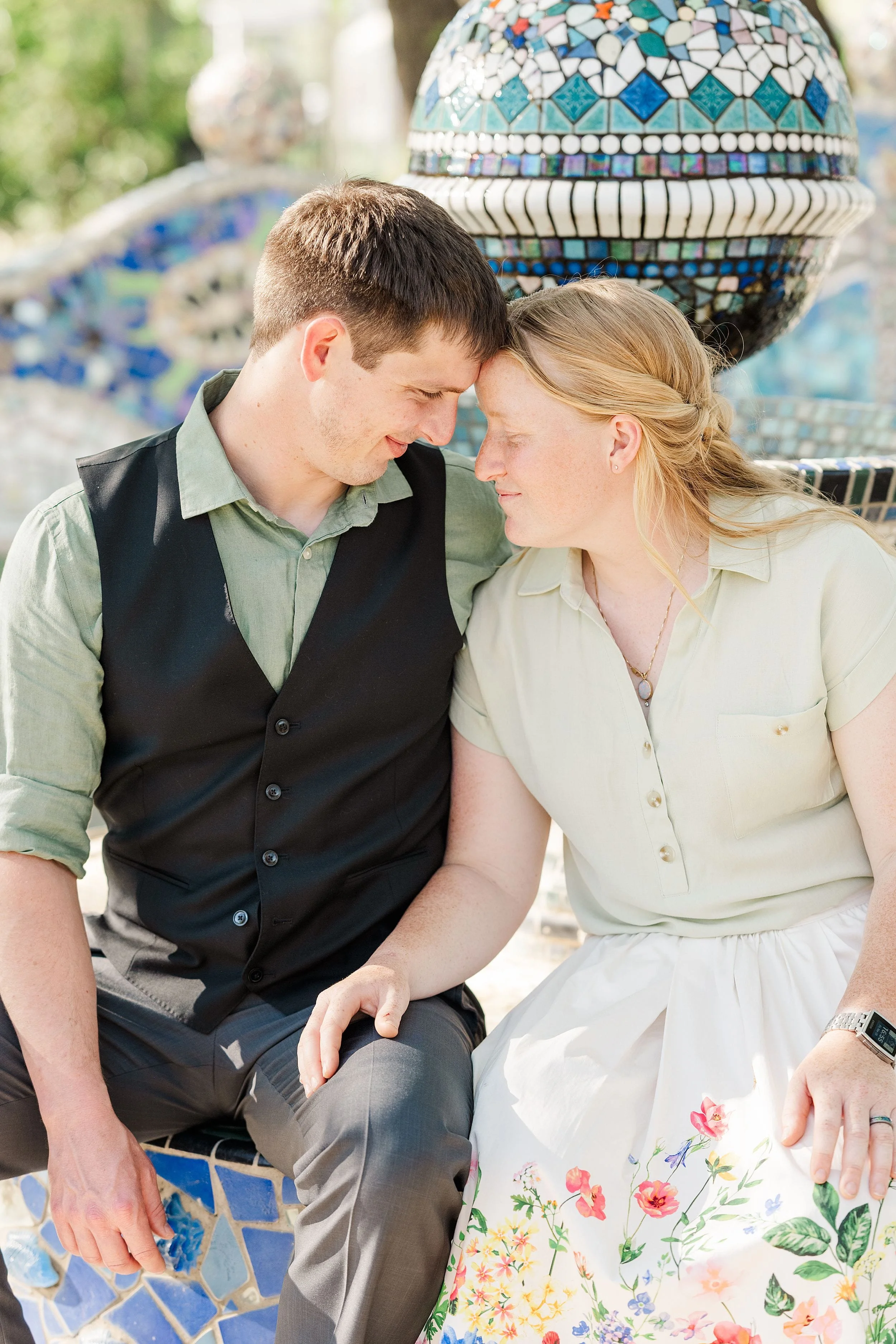 A couple sits closely on a mosaic bench, gently touching foreheads. They wear soft tones, evoking tenderness. A colorful tiled sphere is in the background.