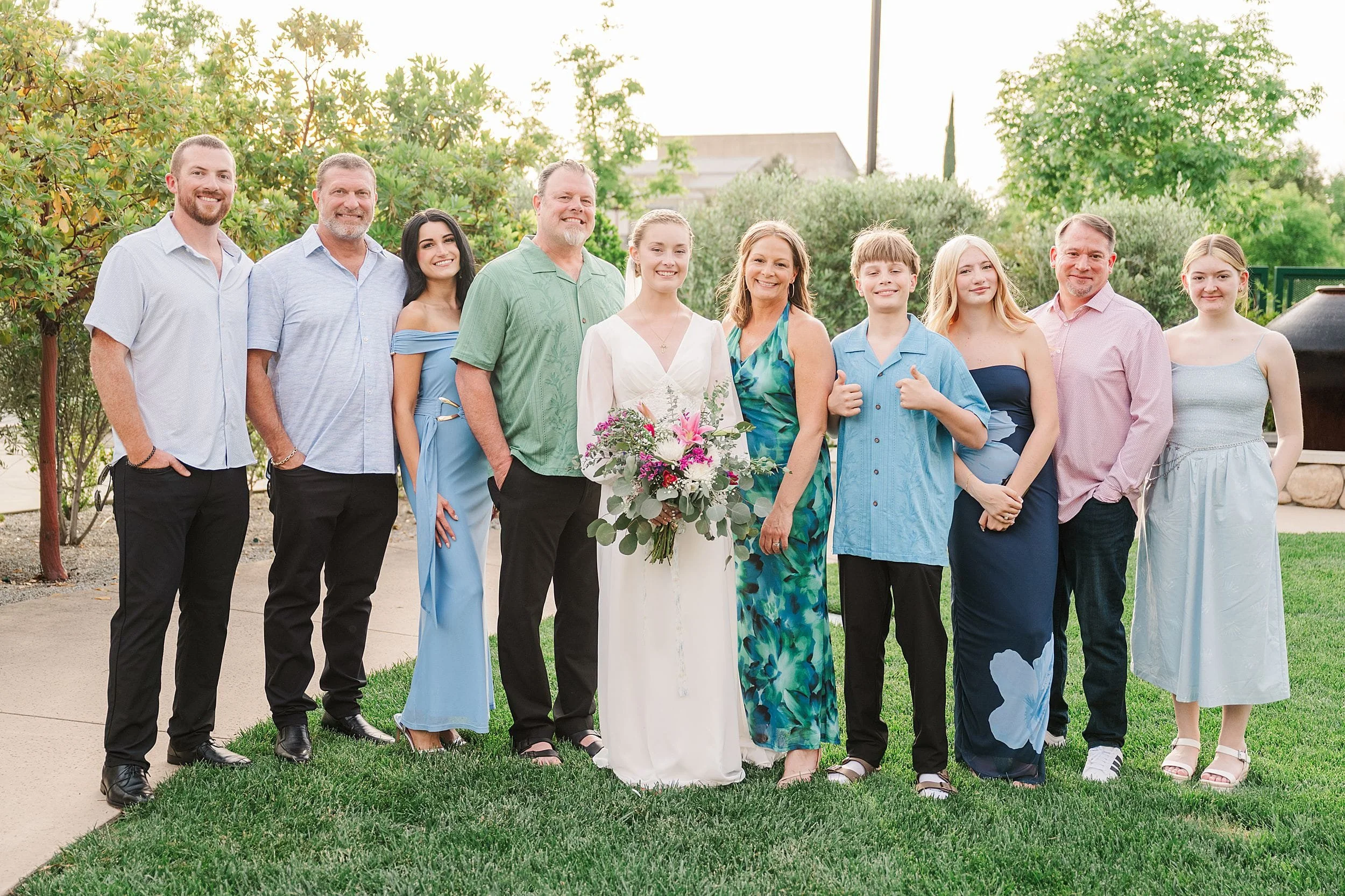 A smiling group poses outdoors in casual and formal attire. A woman in a white dress holds a flower bouquet. Lush greenery and a relaxed atmosphere surround them.