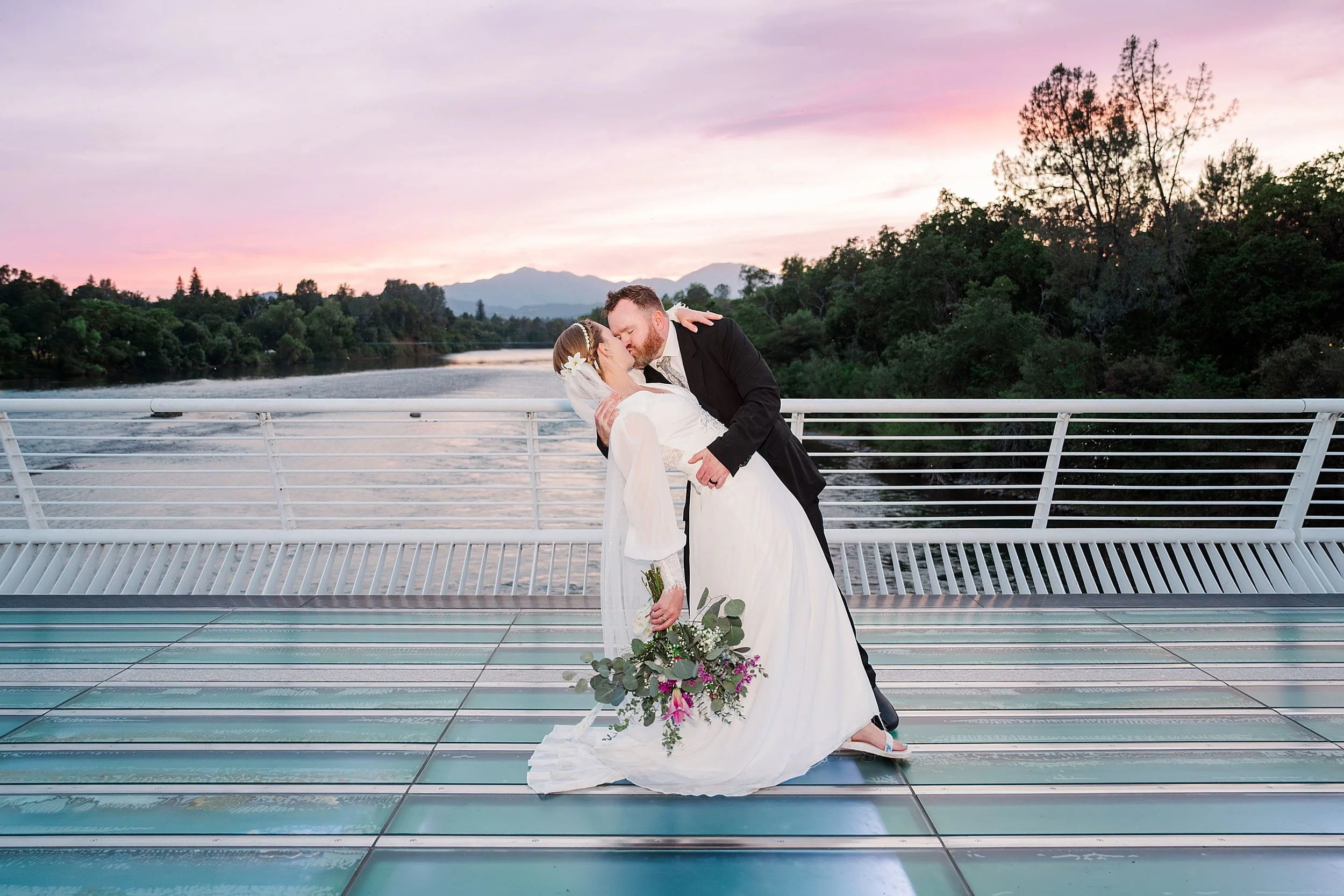 Bride in a flowing white dress, holding flowers, is dipped back by groom in black suit on a bridge. Romantic sunset and river view create a serene backdrop.