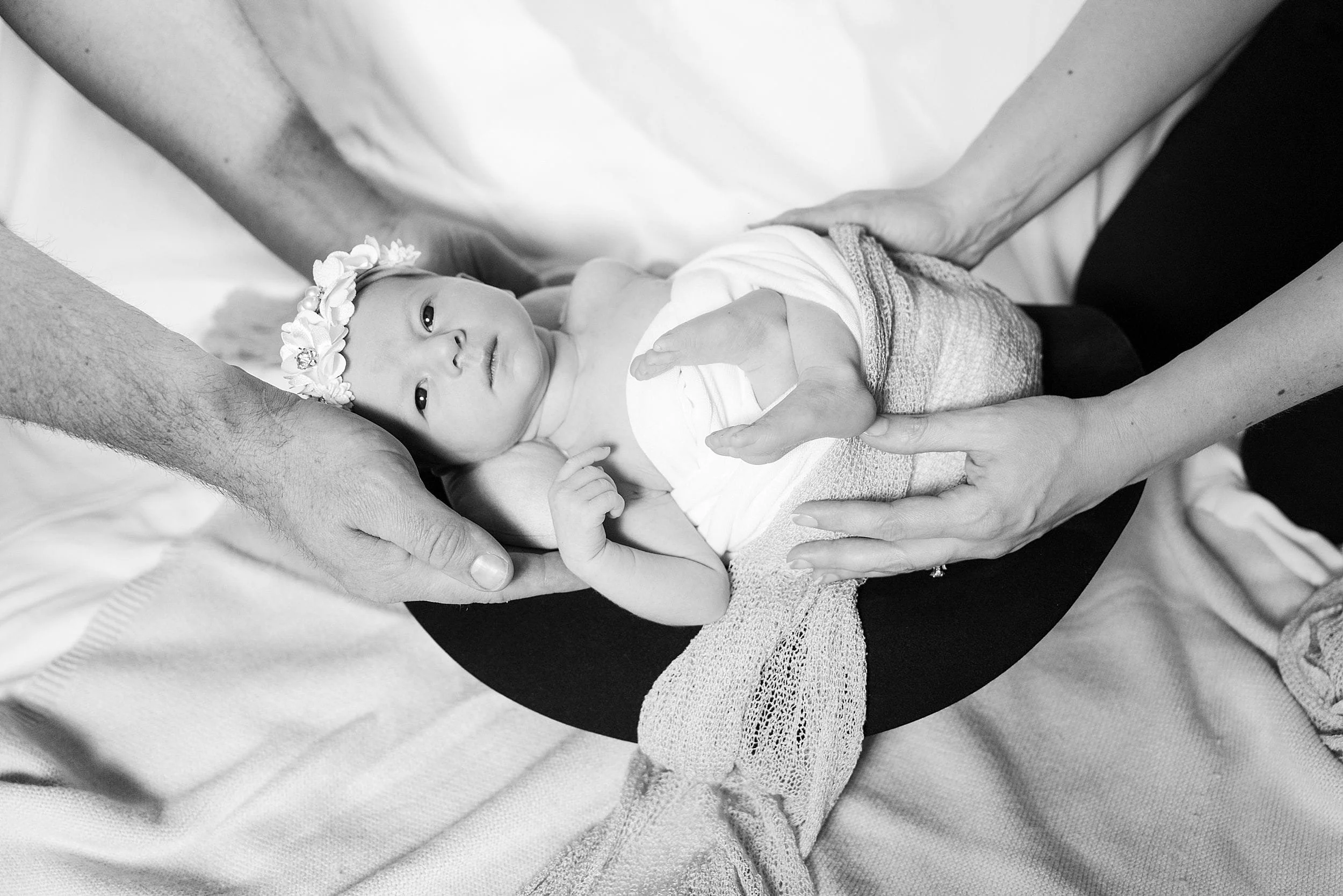 Black and white image of a baby with a floral headband, swaddled in a soft blanket. Two pairs of adult hands gently cradle the baby, conveying warmth and protection.