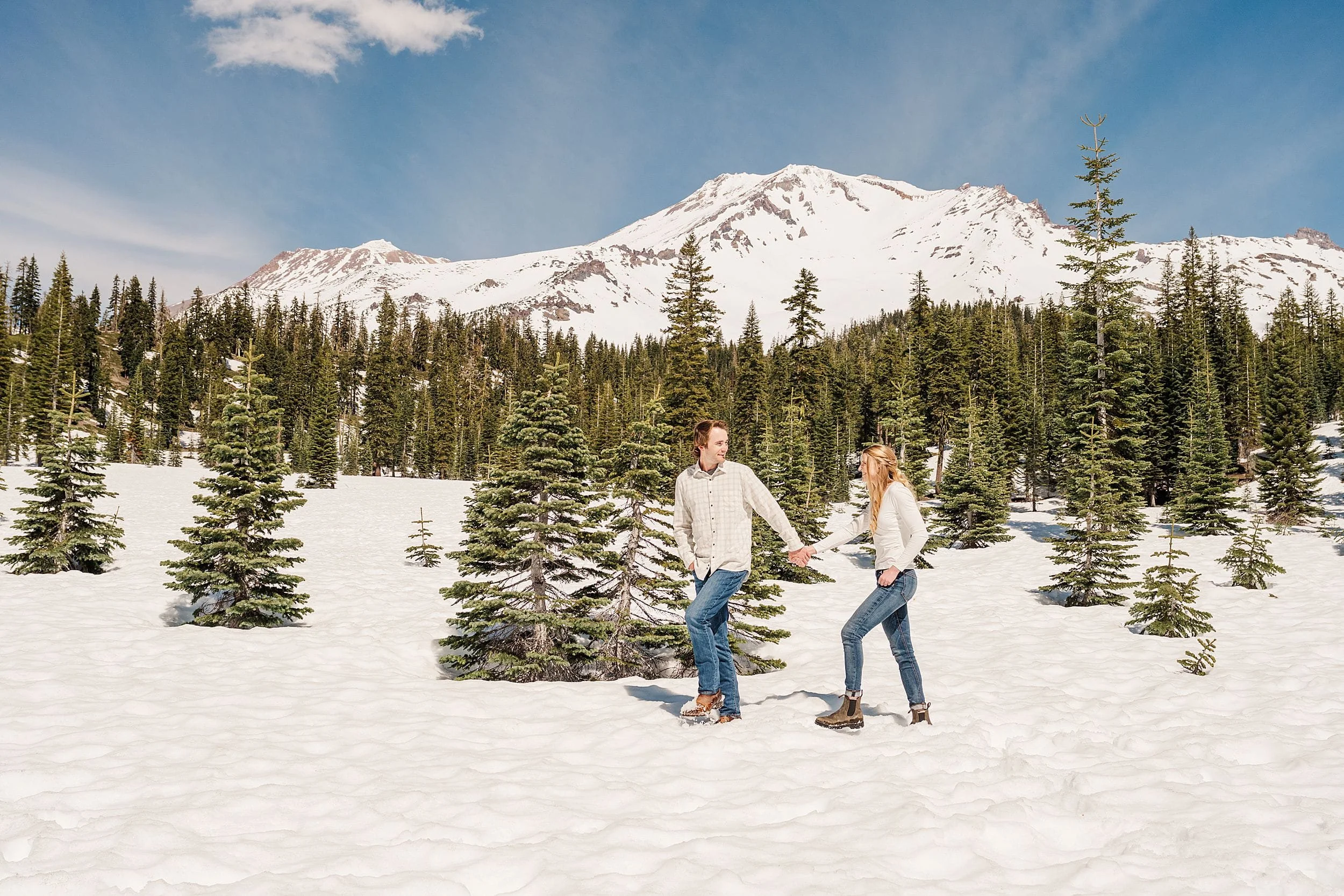 A couple joyfully walks through a snowy landscape with evergreen trees and a snow-capped mountain in the background, under a clear blue sky.