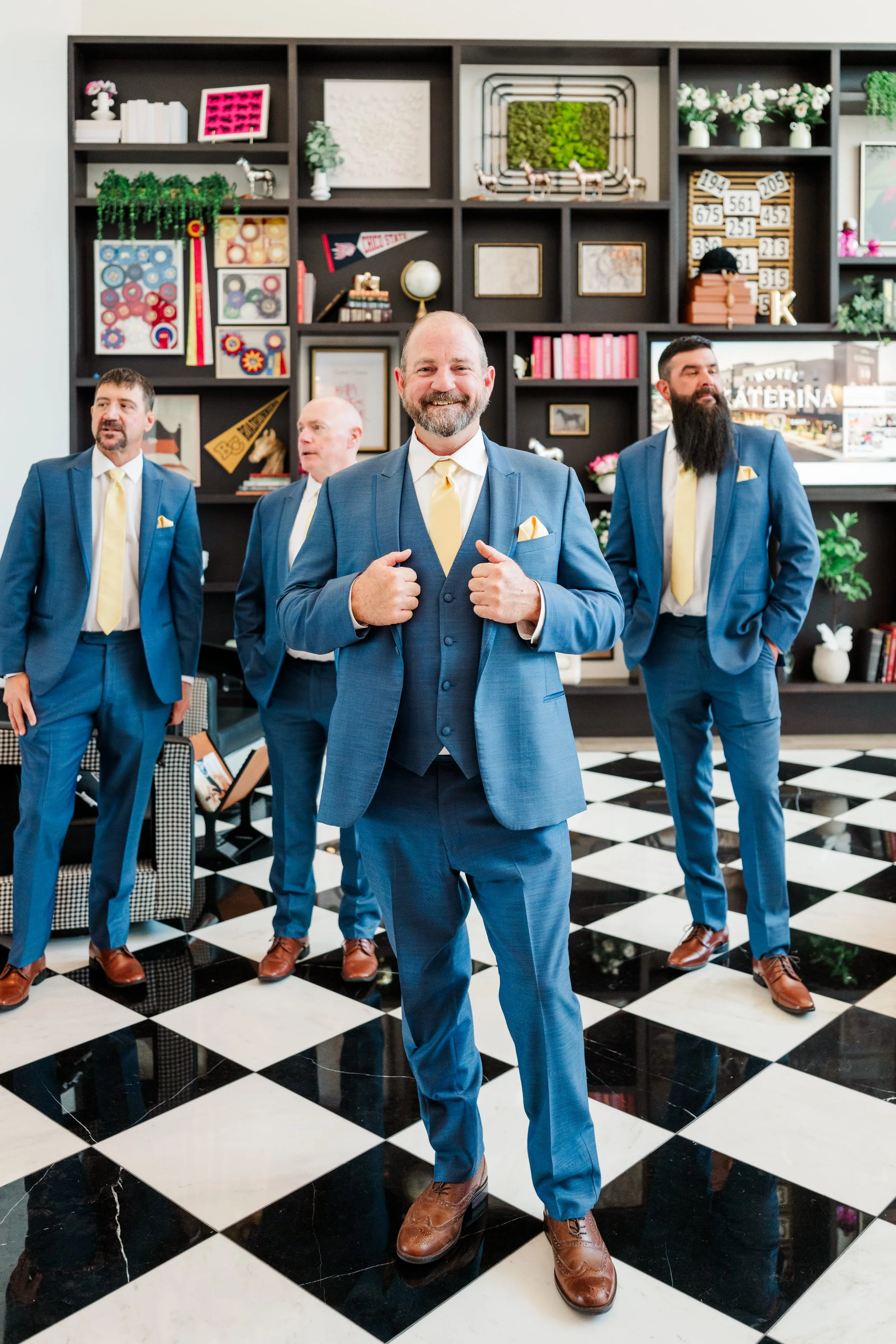 Four men in blue suits with yellow ties and pocket squares standing in a stylish black and white checkered floor room with a black bookshelf decorated with various art and vintage items. The man in the front center is smiling and adjusting his lapels