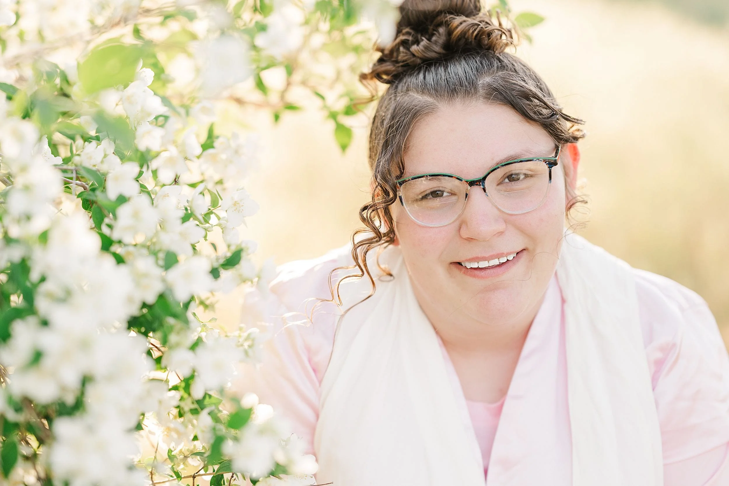 A smiling person with glasses stands near a bush of white flowers in a sunlit garden. They wear a light pink shirt and scarf. The scene is bright and serene.