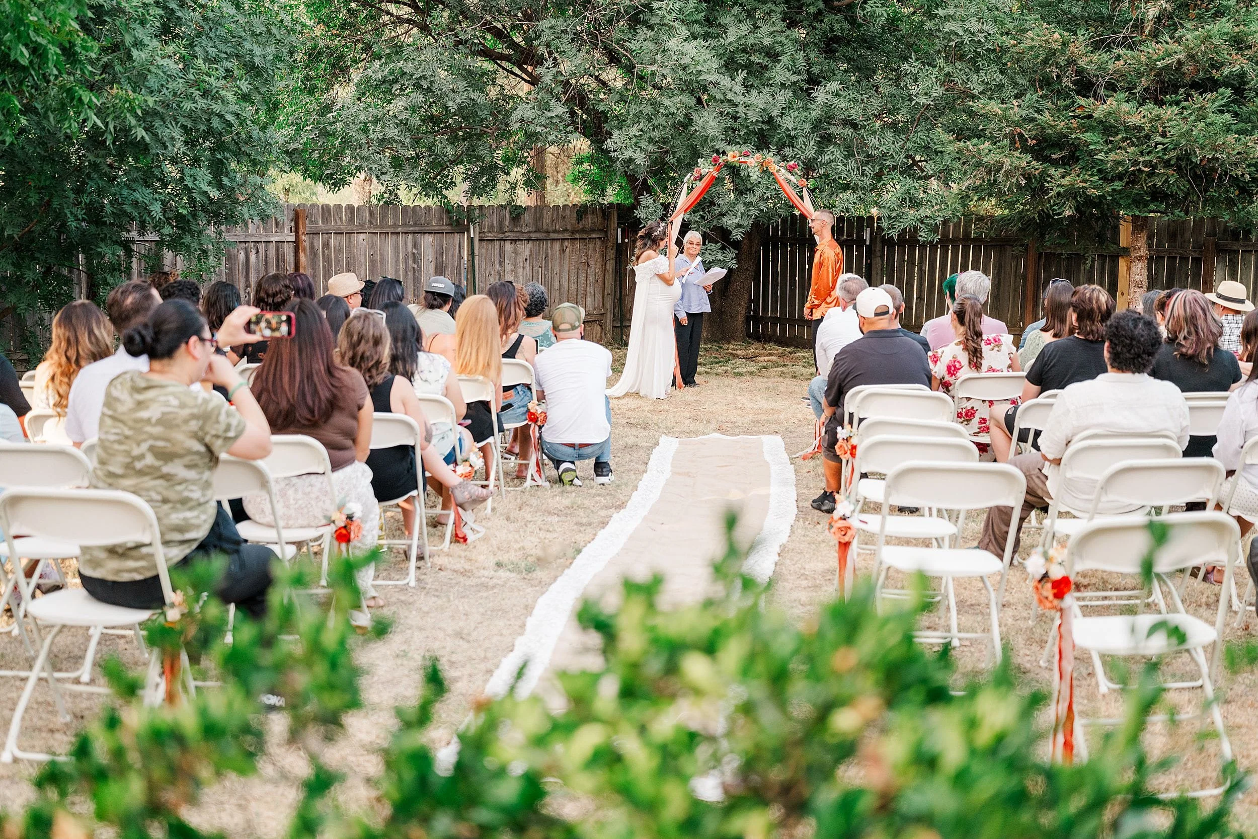 Outdoor wedding ceremony in a leafy garden, with guests seated on white chairs as the couple stands beneath a colorful floral arch.