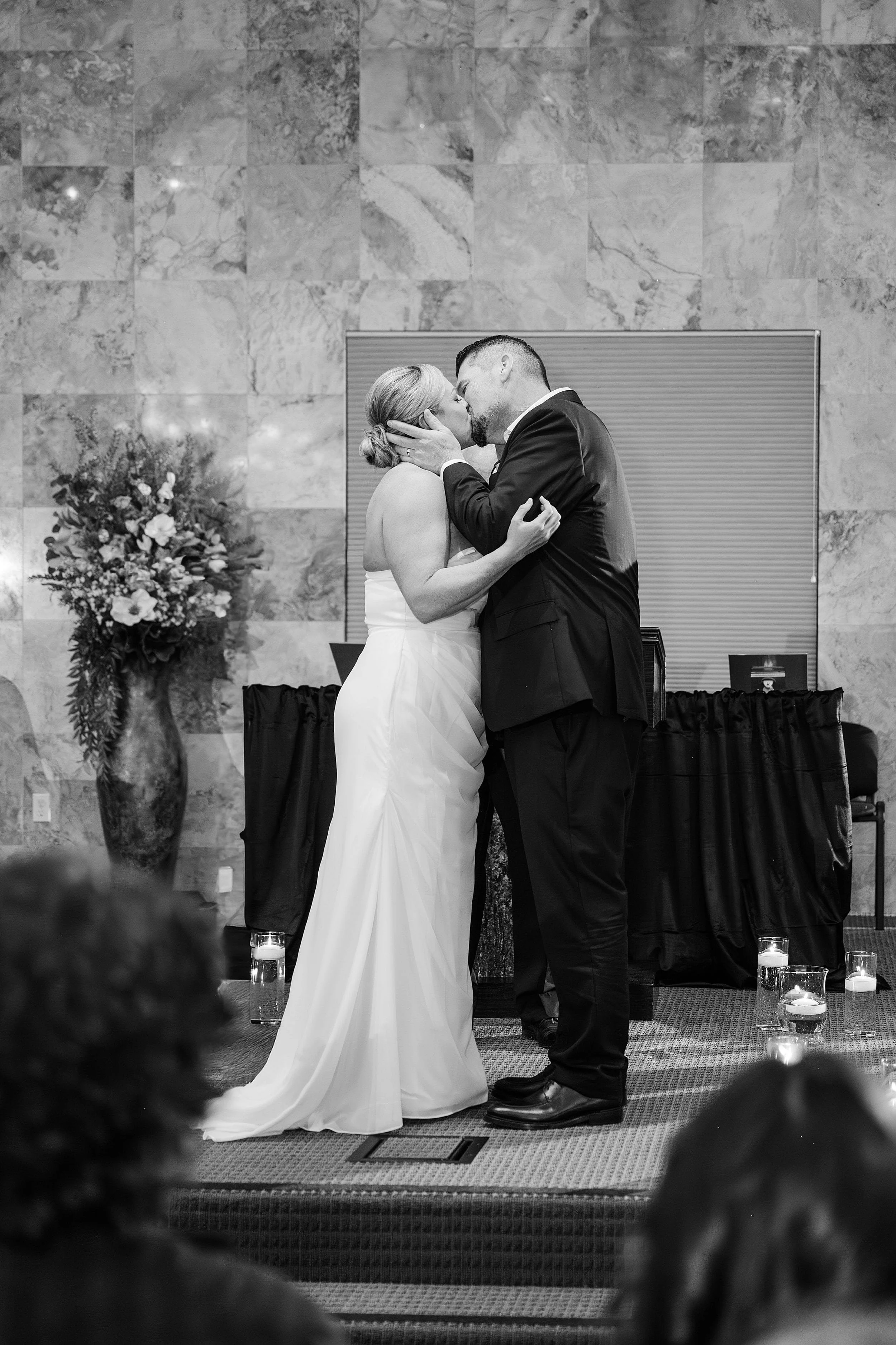 A newlywed couple shares a kiss at the altar, surrounded by candles and flowers in a vase, conveying joy and intimacy in a formal setting.