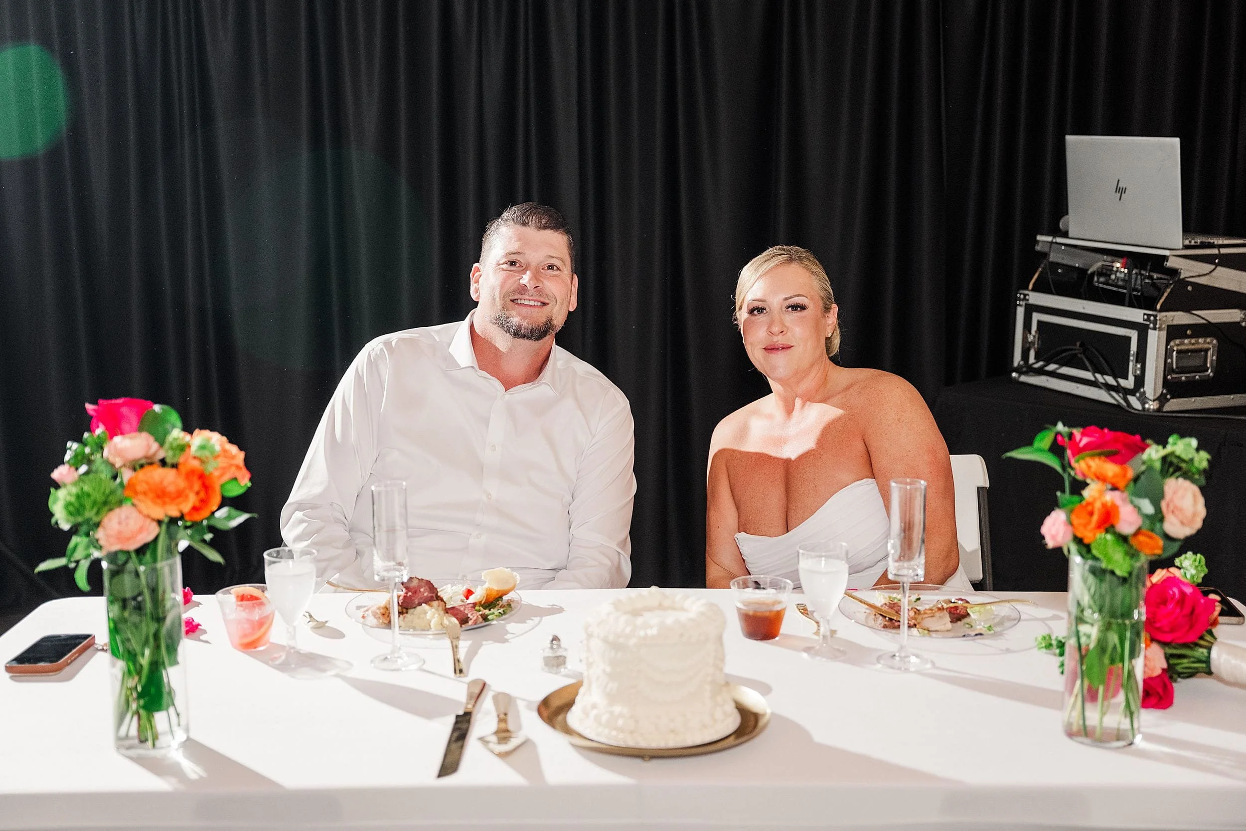 A couple sits at a table dressed in wedding attire, smiling. A white cake and colorful flowers adorn the table. The atmosphere is joyful and celebratory.