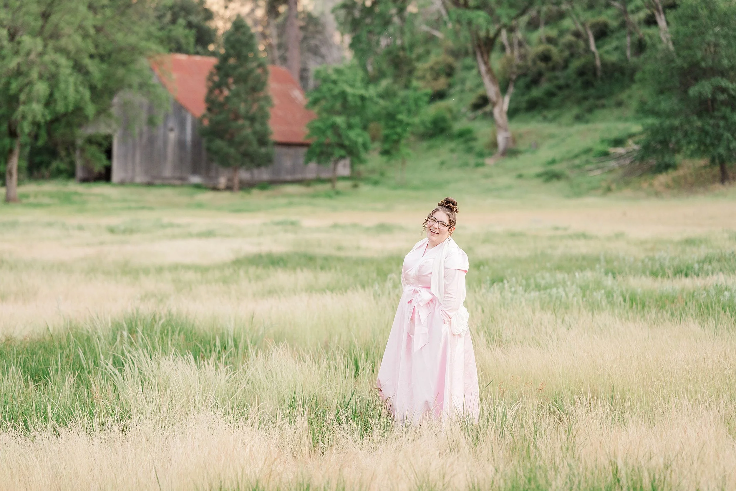 Woman in a pink dress stands in a lush green field with a soft smile. A rustic barn with a red roof is in the background under a cloudy sky.